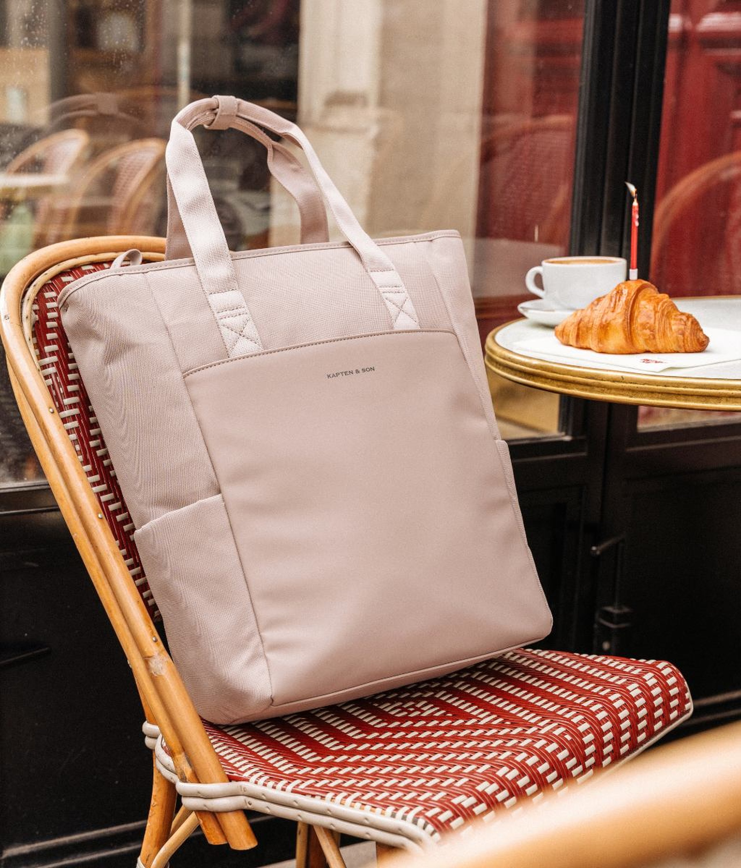 Große hellrosa Kapten & Son Wickeltasche mit Tragegriffen und Seitentaschen, auf einem roten Korbstuhl in einem Pariser Café, mit einem Croissant, einer Kaffeetasse und einer brennenden Geburtstagskerze auf einem Bistrotisch im Hintergrund.