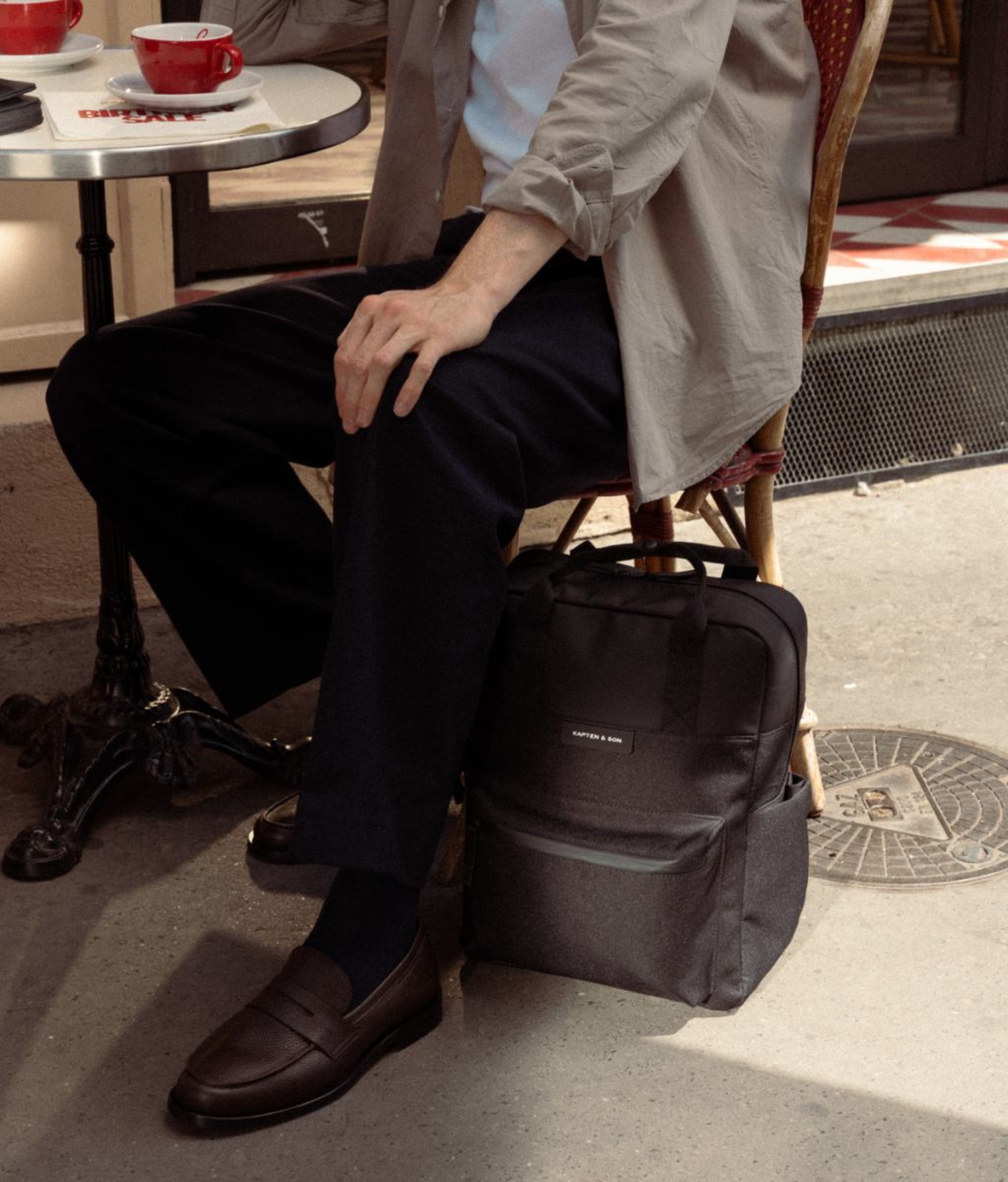 Man sitting on a wicker café chair in a Parisian café, wearing dark trousers, a light grey jacket and dark brown loafers, with a dark grey Kapten & Son backpack with top handles placed on the ground beside him and a red coffee cup visible on the bistro table in the background.