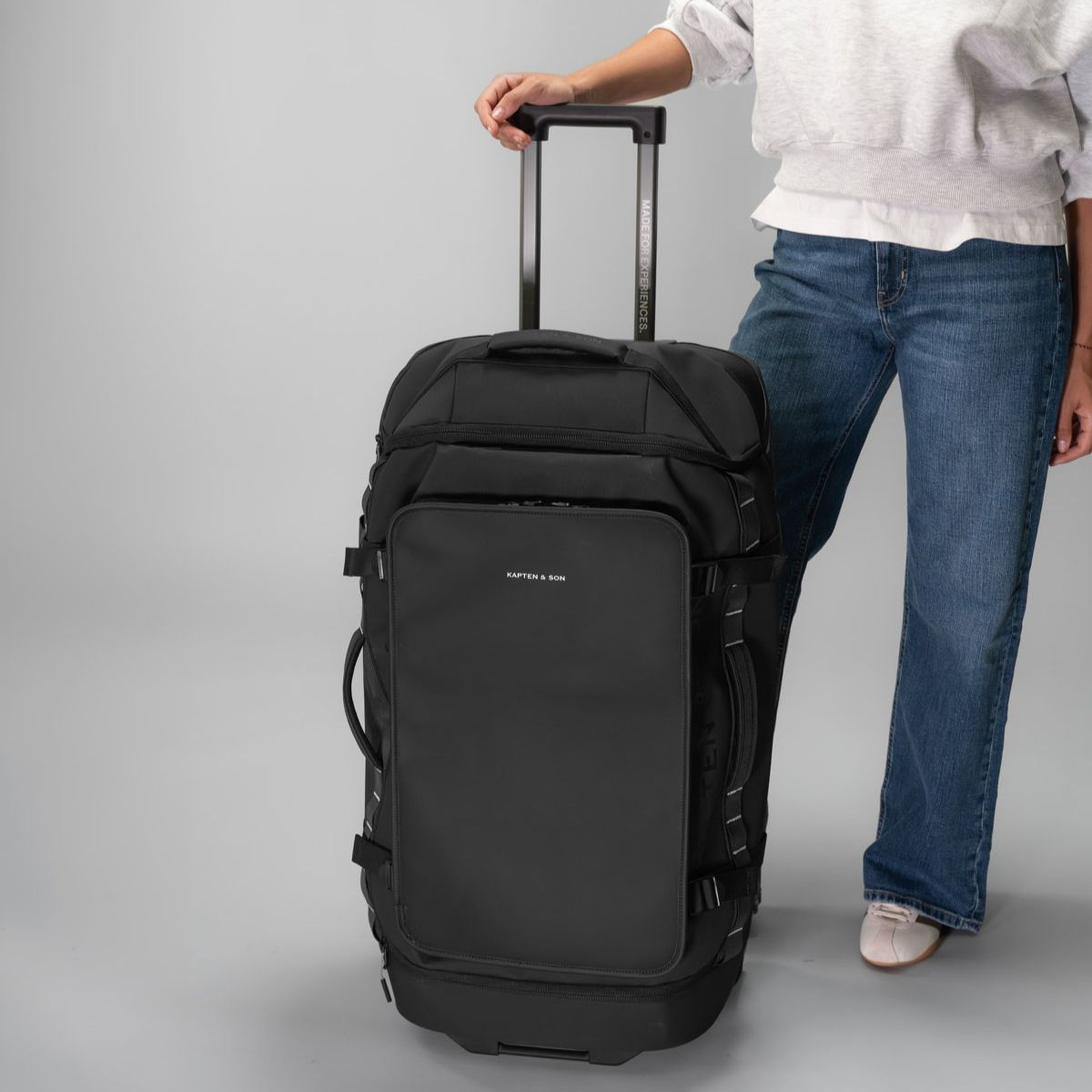 Person in jeans and white sneakers holding a large black Kapten & Son suitcase with an extended handle against a gray background.