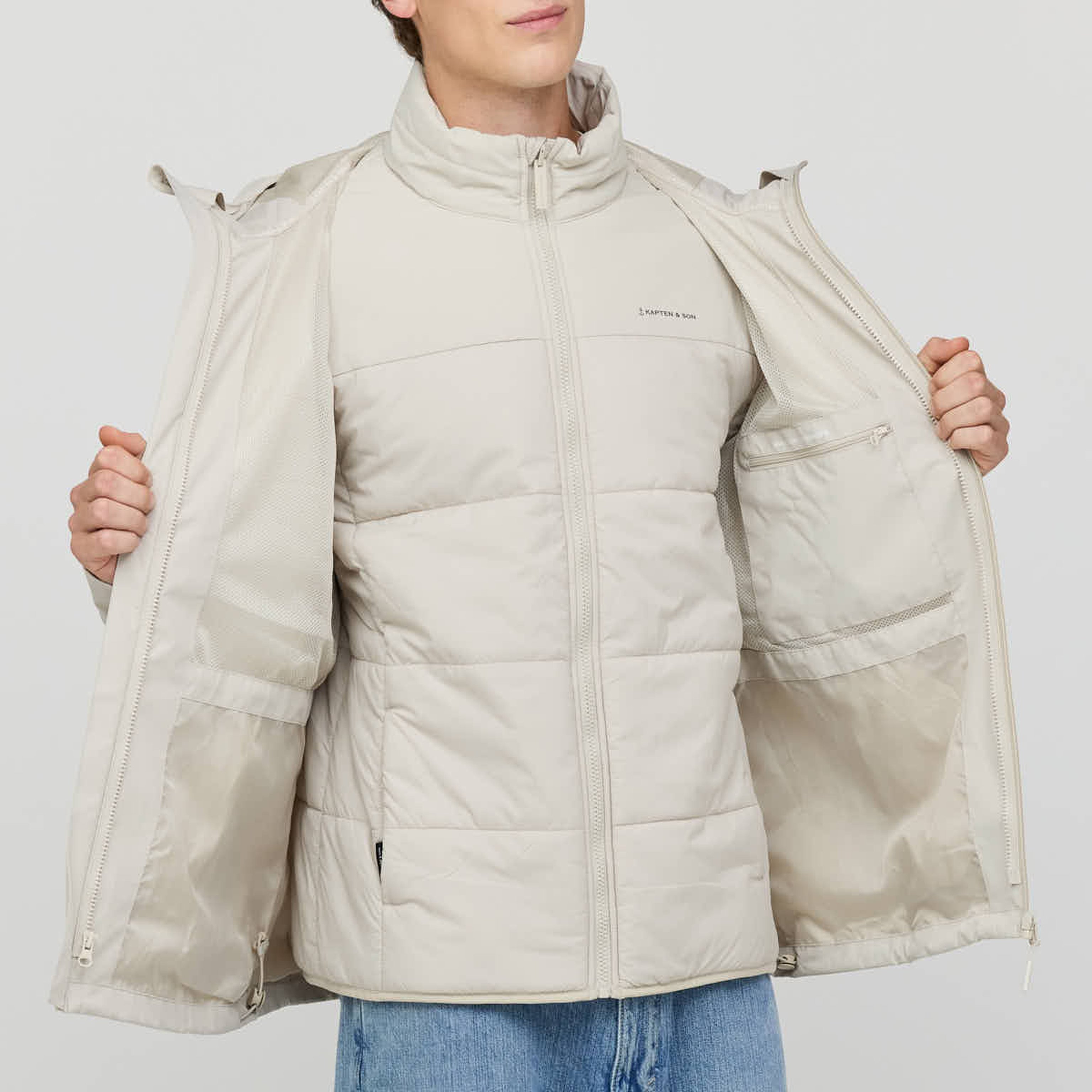 Eine Person in einer hellbeigen, gesteppten Jacke, die vorne offen (unzippend) getragen wird, steht vor einem schlichten weißen Hintergrund. Sie trägt blaue Jeans und hat einen selbstbewussten Ausdruck.