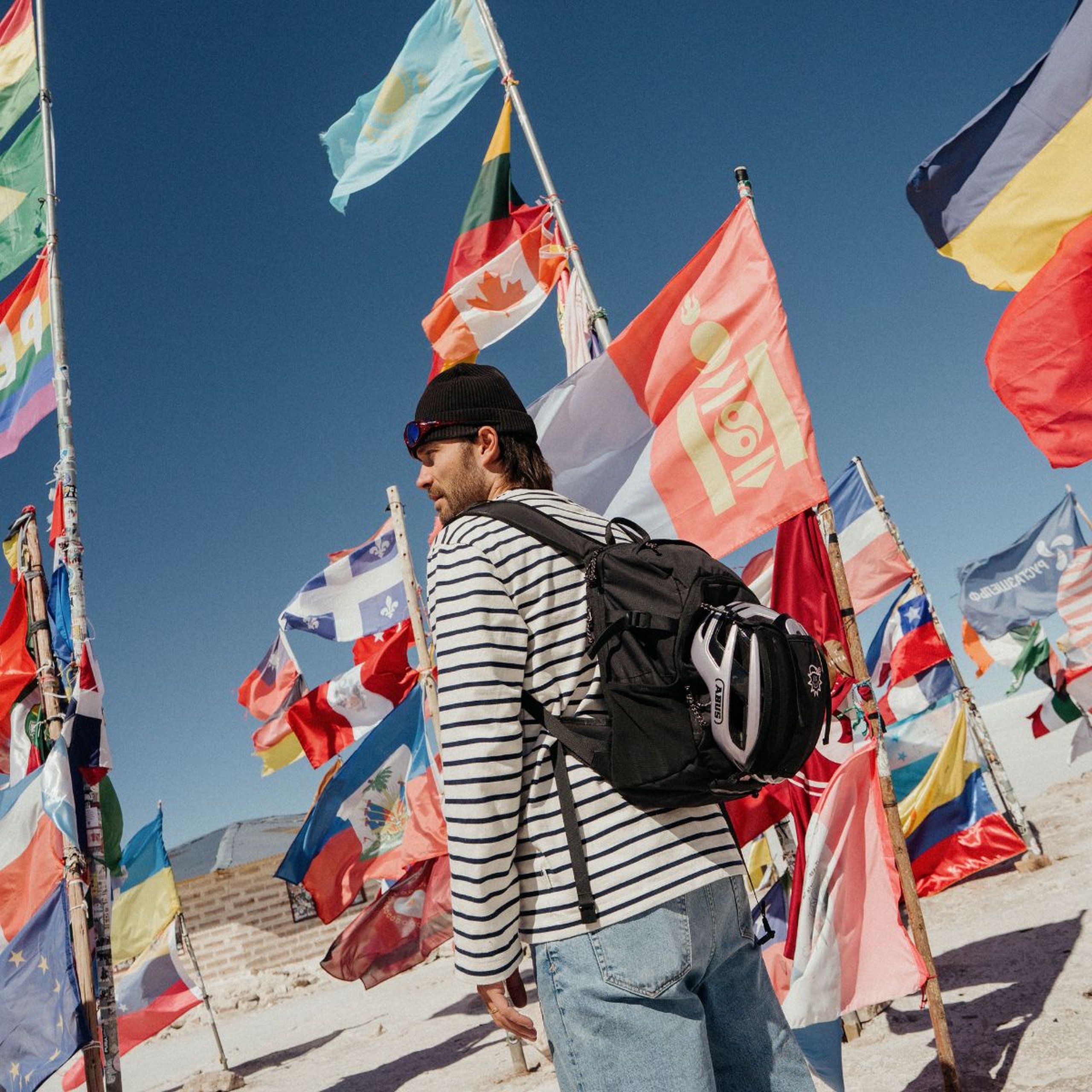 Person mit gestreiftem Langarmshirt, Beanie und Rucksack, umgeben von zahlreichen internationalen Fahnen unter einem strahlend blauen Himmel.