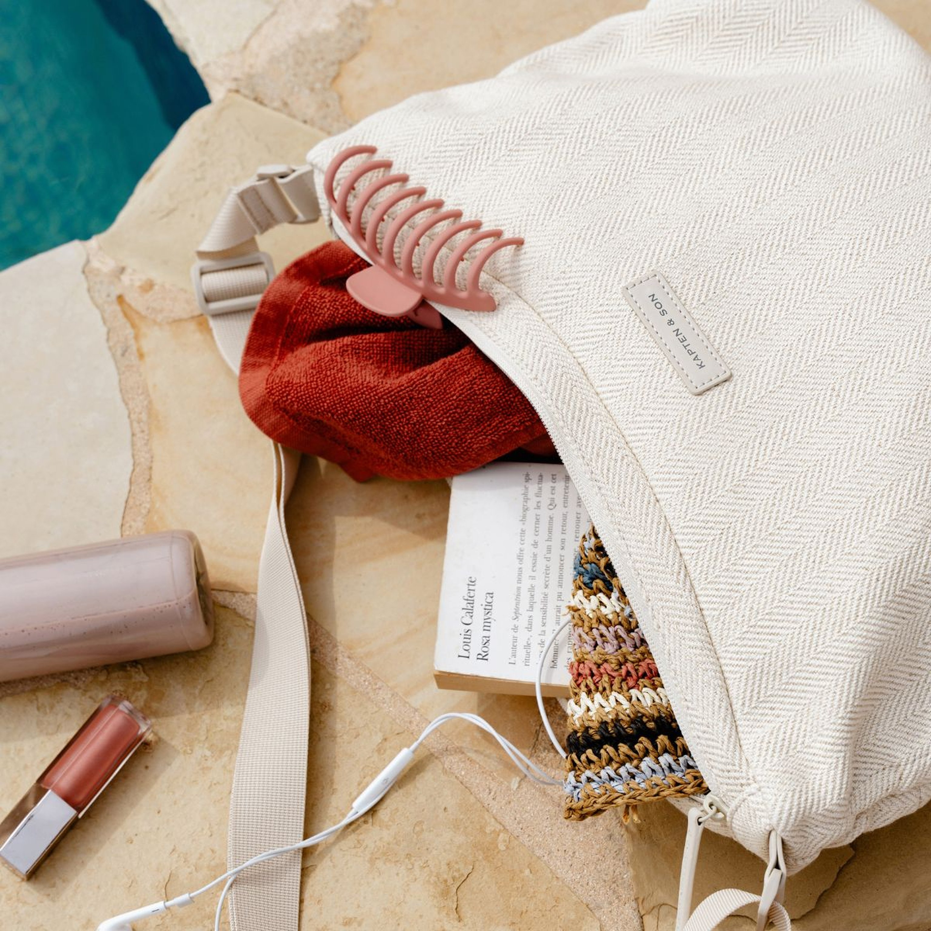 Un sac beige clair est ouvert sur une surface en pierre au bord d'une piscine, laissant apparaître une serviette rouge, un livre et un article en maille. À proximité se trouvent une bouteille et un gloss, dans une atmosphère calme et détendue.