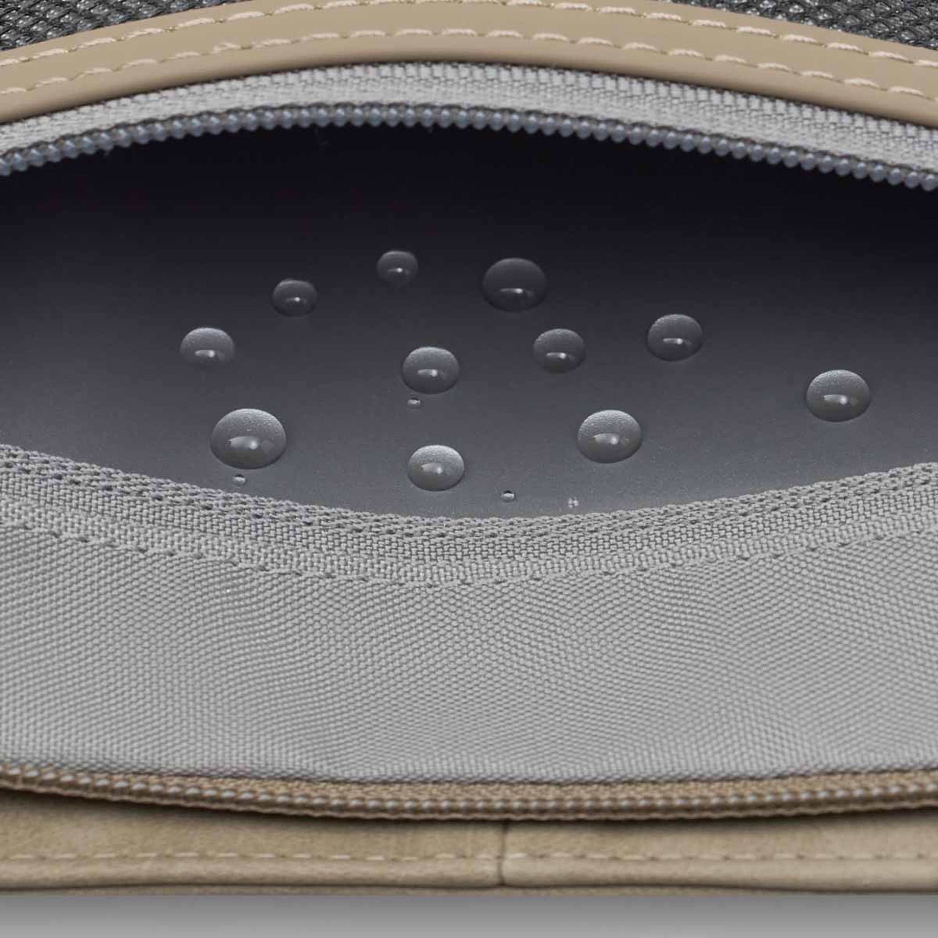 Close-up of water droplets on a gray fabric surface inside an open bag with visible stitching and a slightly curved zipper, conveying water-resistance.