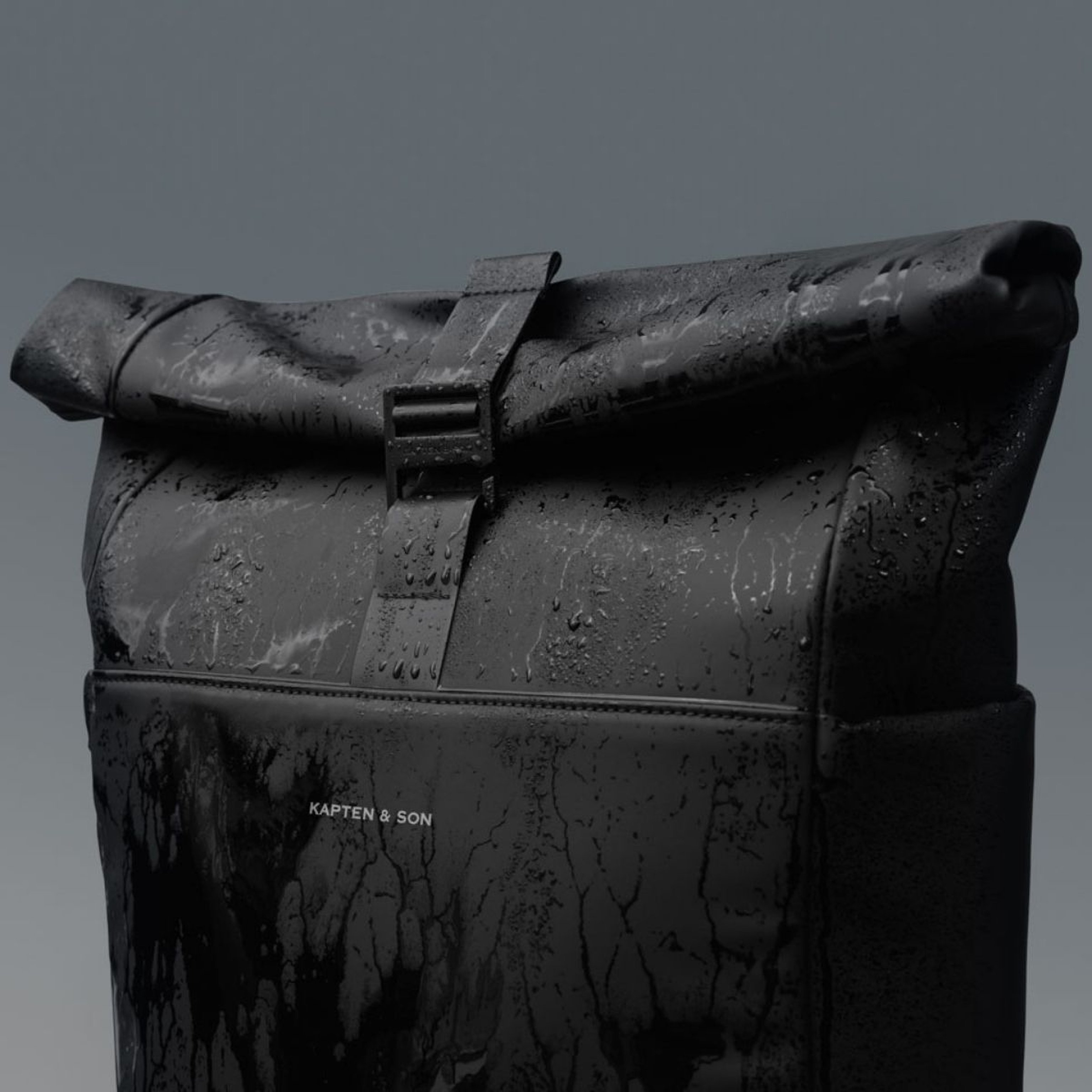 Sleek black backpack with a roll-top design, covered in raindrops. The brand name "Kapten & Son" is visible, conveying elegance and durability.