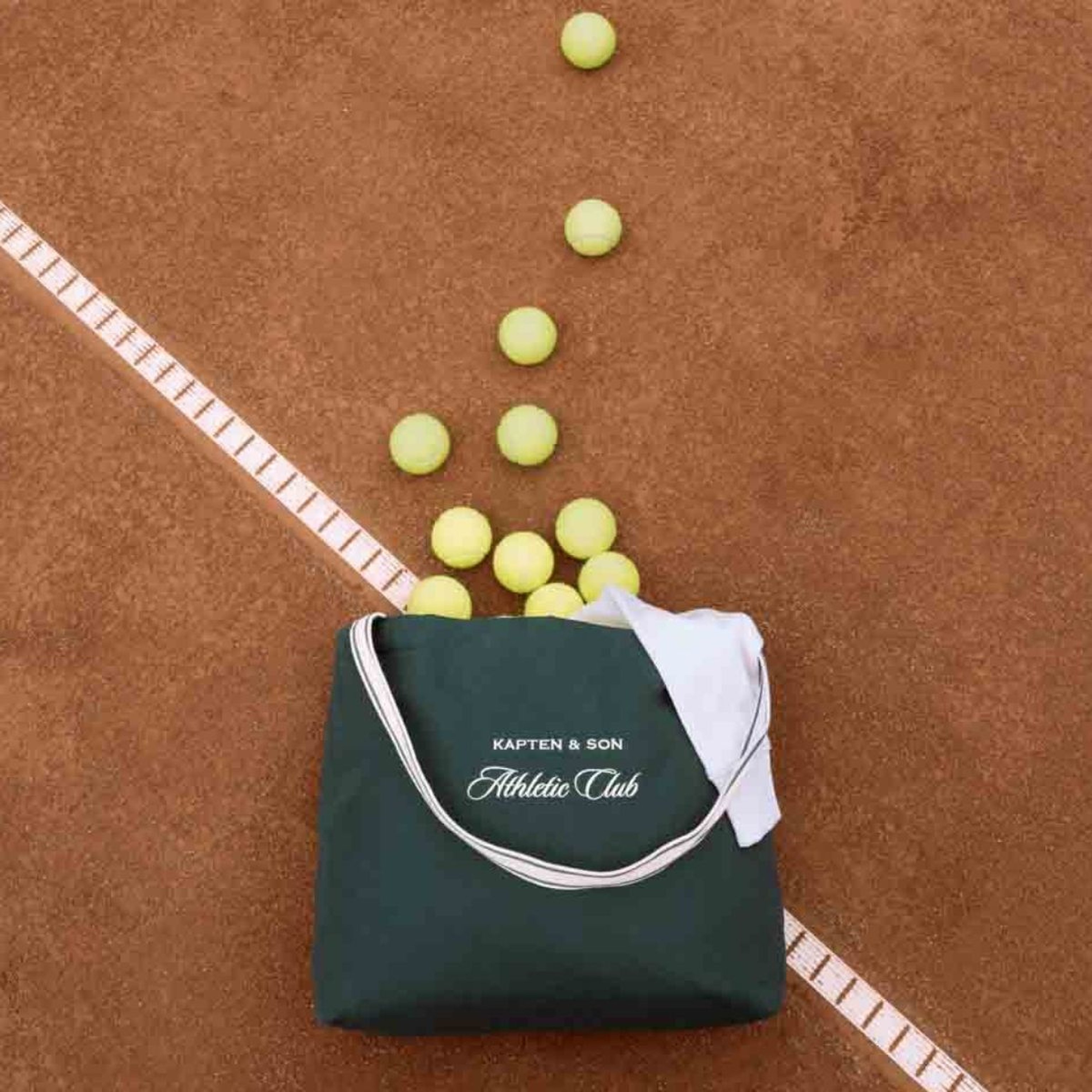 A green bag labelled "Kapten & Son Athletic Club" spills yellow tennis balls onto a clay tennis court, with a white line marking the boundary.