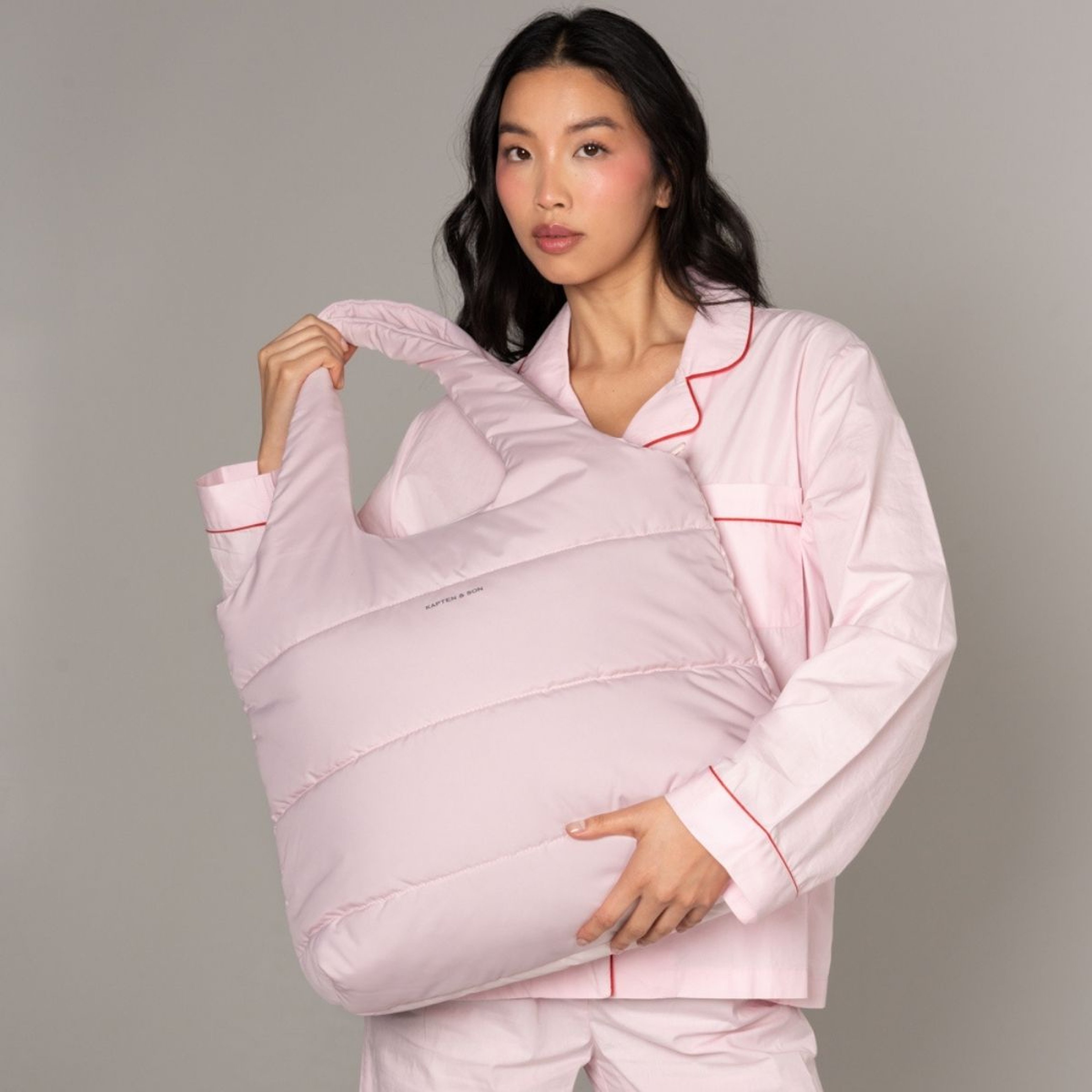 Eine Frau in hellrosa Pyjama hält eine passende, oversized gesteppte Tasche. Die Szene vermittelt eine cozy, entspannte Stimmung mit weichen, beruhigenden Farbtönen.