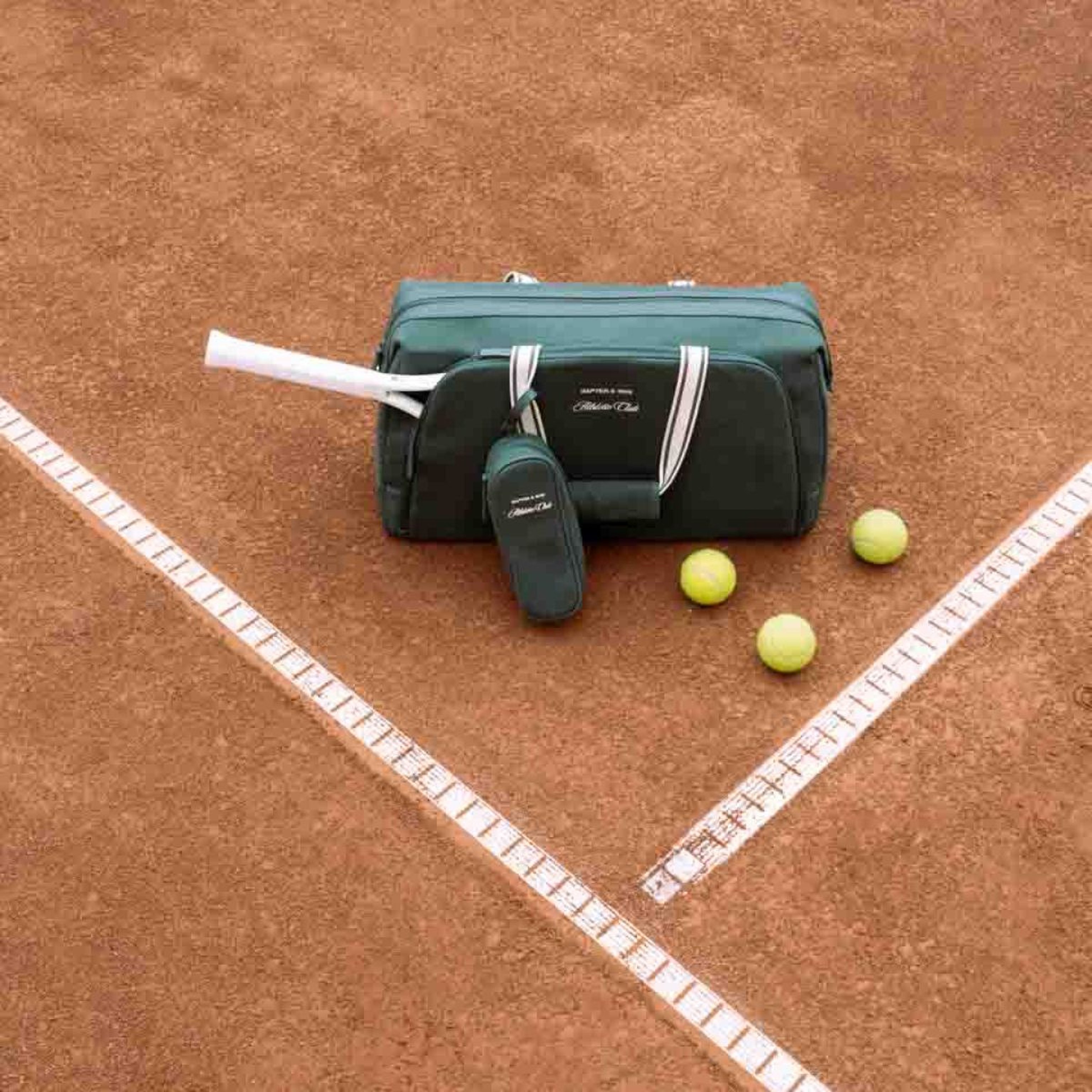 Eine grüne Tasche mit der Aufschrift „Kapten & Son Athletic Club" lässt gelbe Tennisbälle auf einen Sandplatz fallen, mit einer weißen Linie, die die Begrenzung markiert.