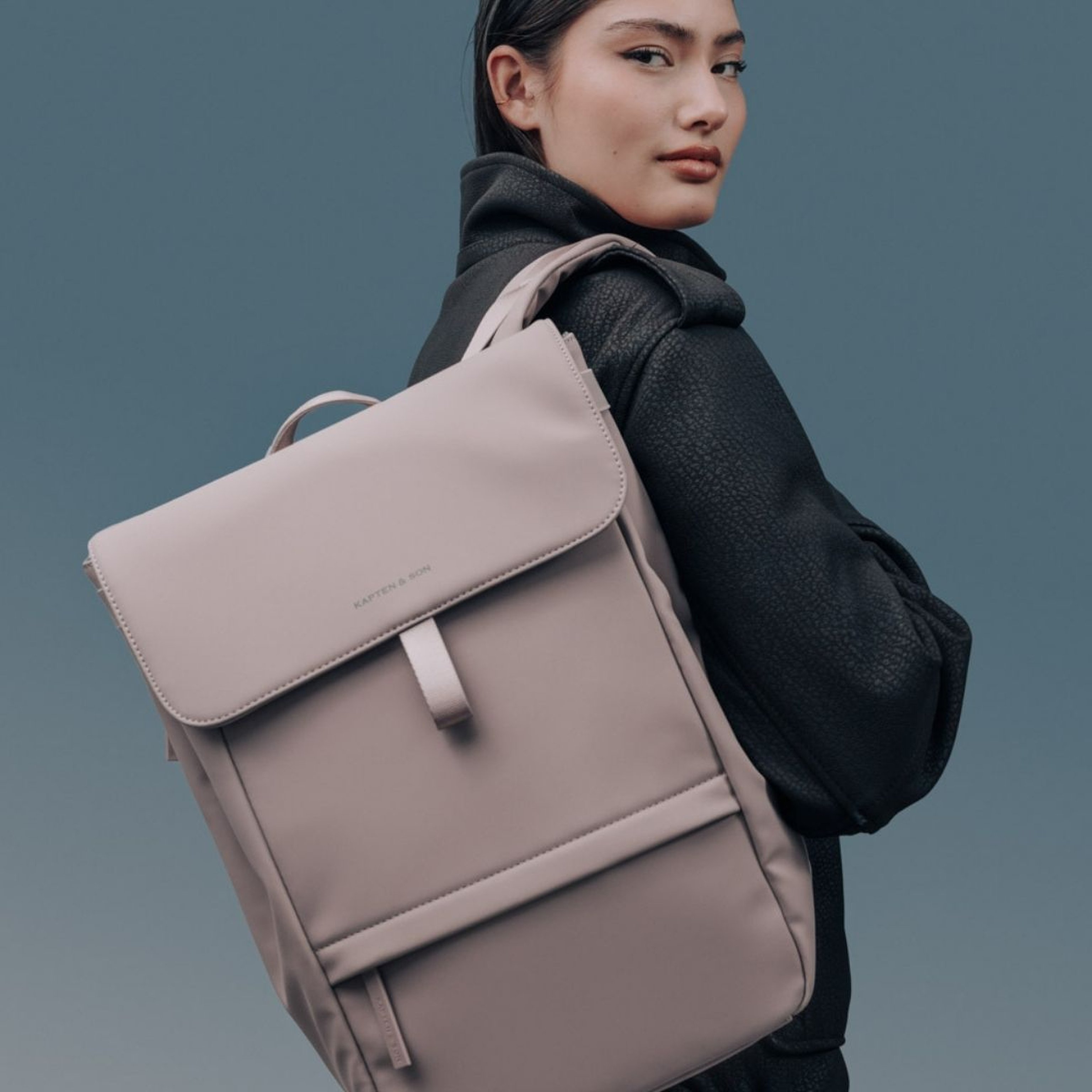 A woman in a black coat poses with a sleek beige backpack against a blue-gray background. Her expression is confident and composed.