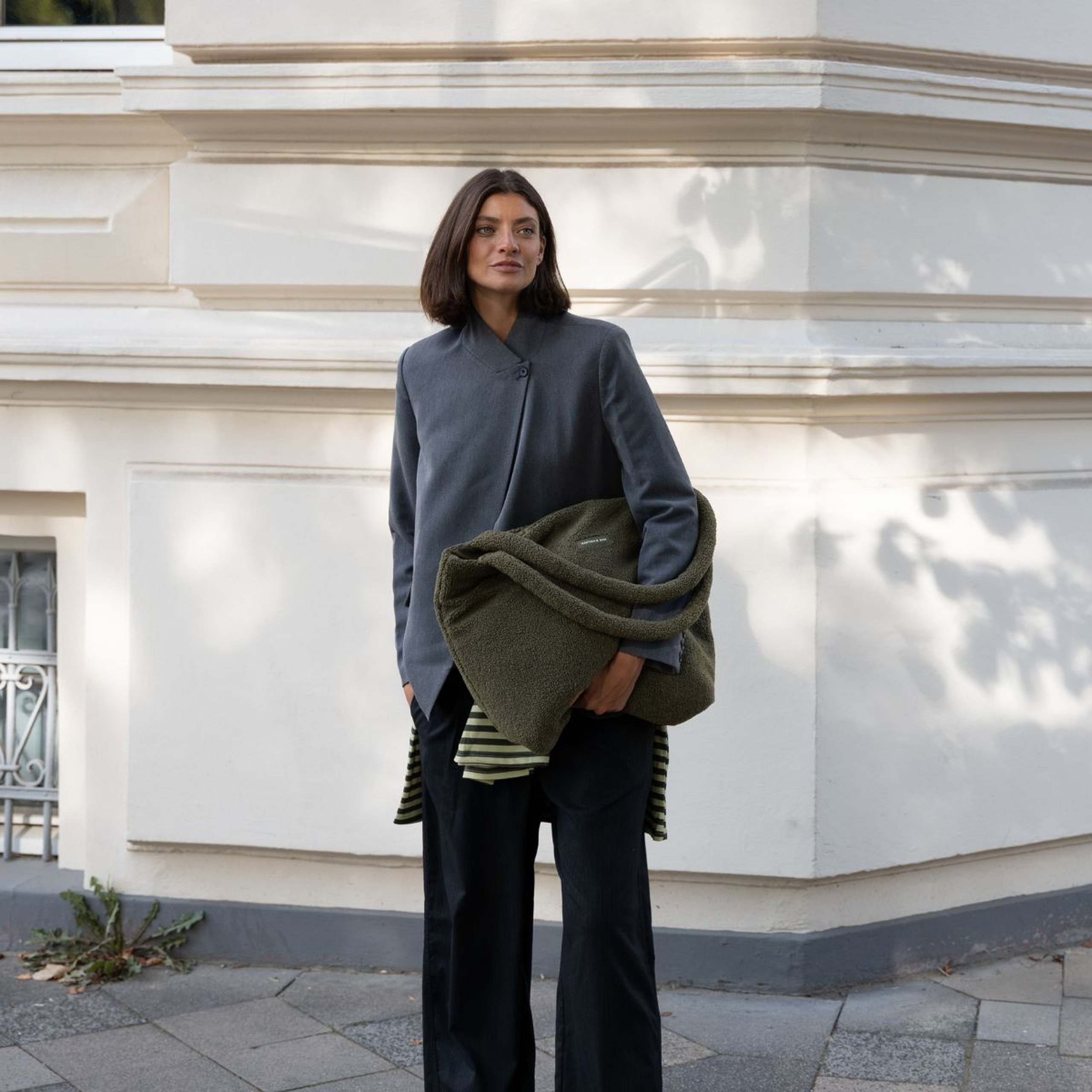 Frau im Blazer hält eine große Kapten & Son Tasche aus weichem Fleece unter dem Arm und steht vor einer Hausfassade.
