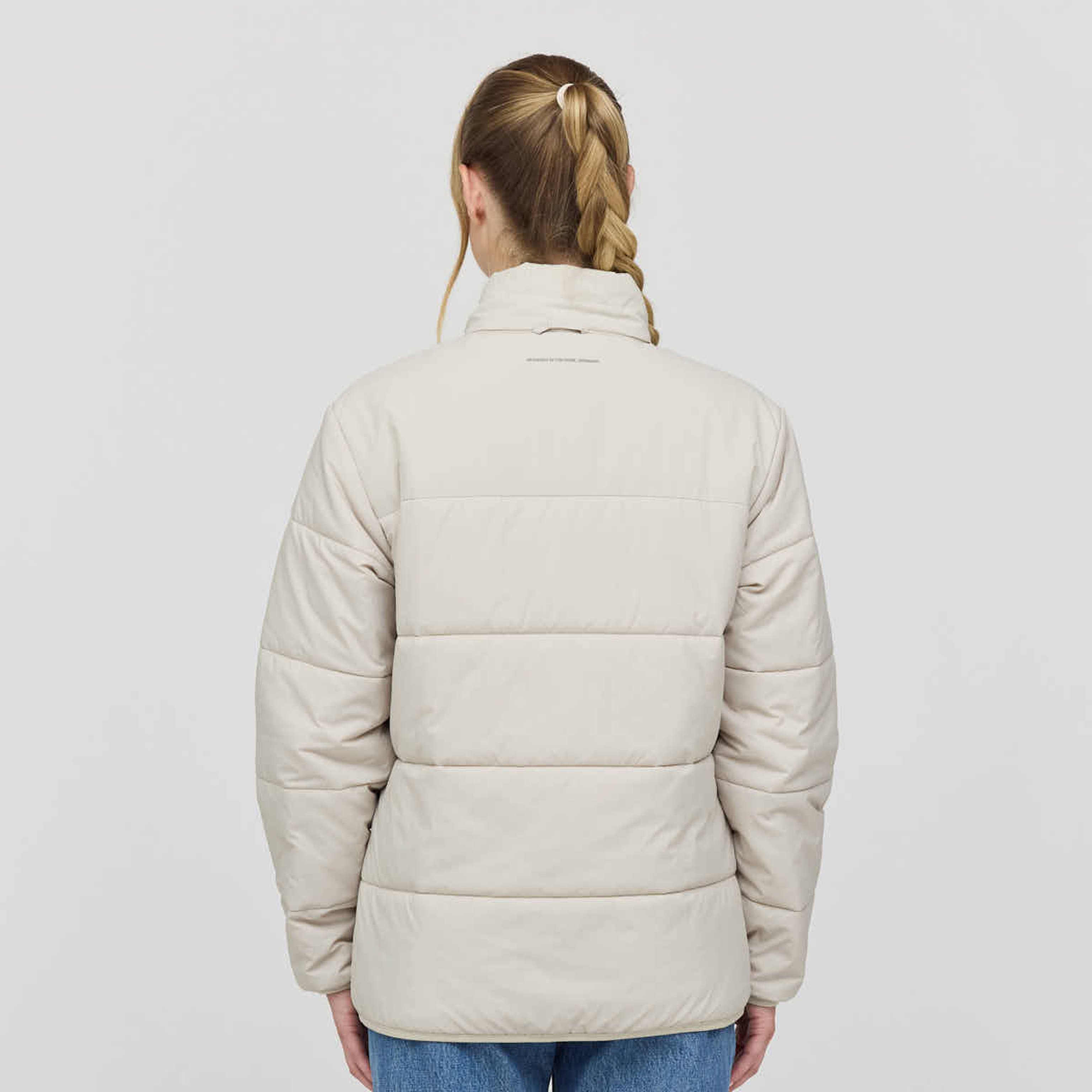 Rear view of a person with a braided hairstyle wearing a beige quilted jacket and blue jeans, standing against a plain white background.