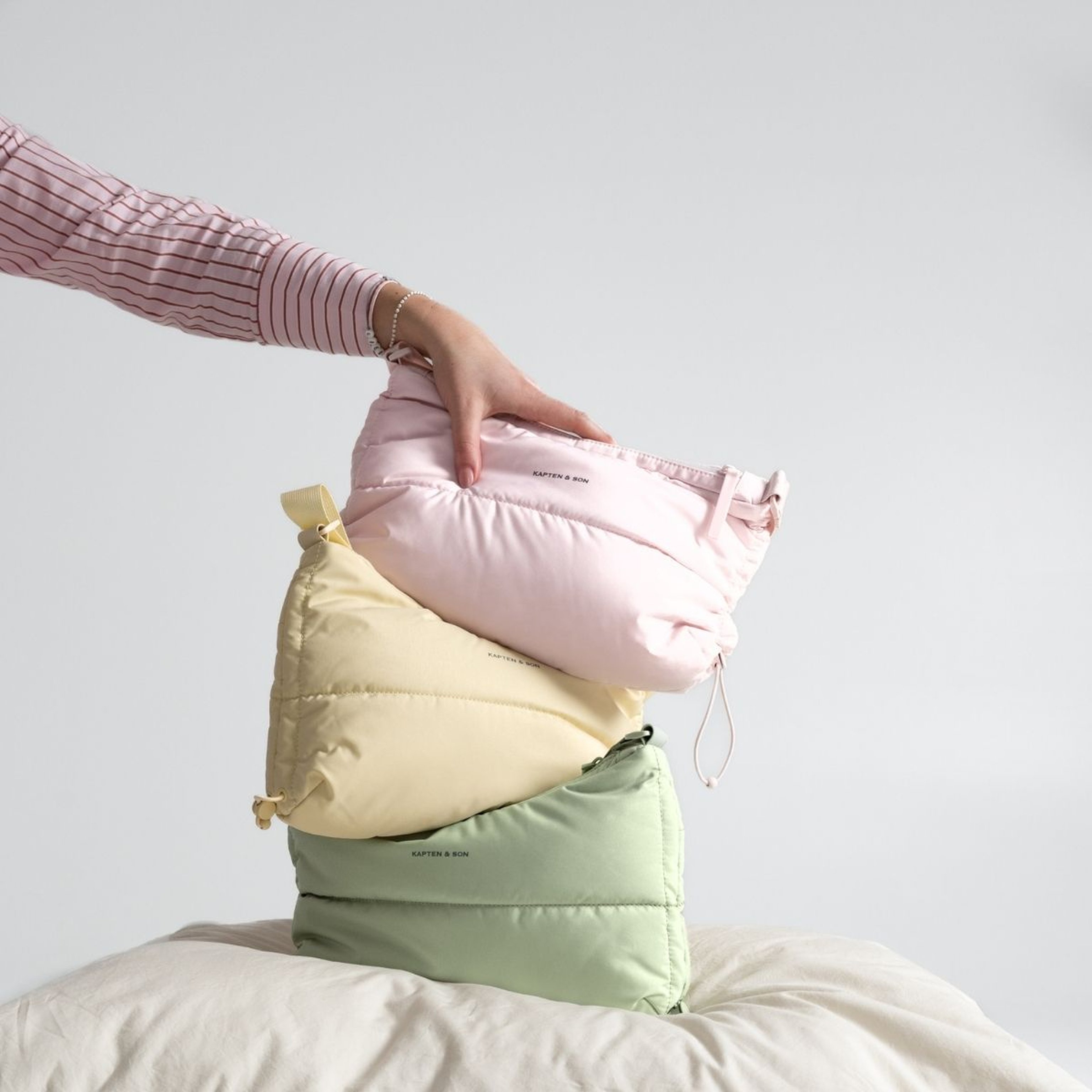 Eine Hand arrangiert drei pastellfarbene Puffer-Bags in Rosa, Gelb und Grün auf einer cremefarbenen Oberfläche. Das Setting ist hell und minimalistisch und vermittelt ein ordentliches, aufgeräumtes Gefühl.