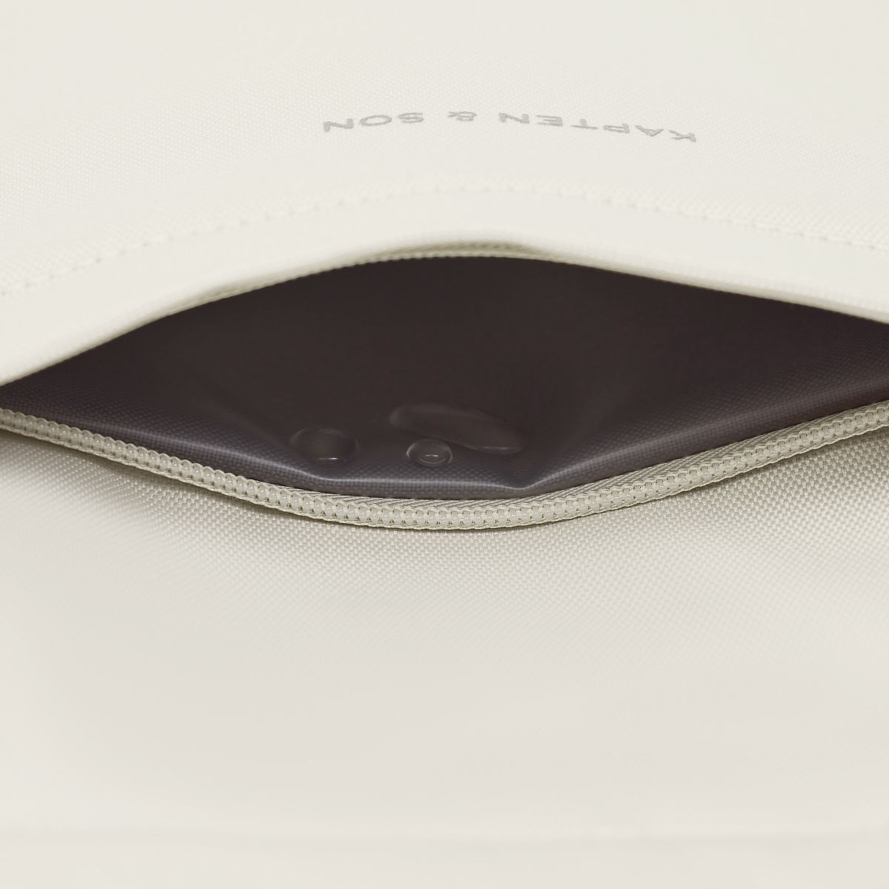 A close-up of a beige fabric bag with a partially open zipper, revealing a black inner lining with two droplets of water, conveying a sense of waterproof protection.