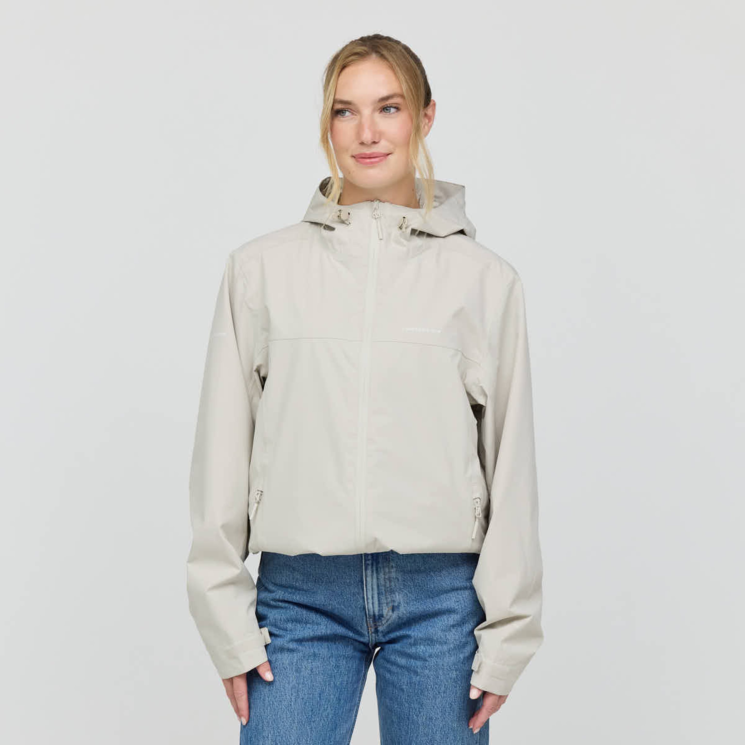 Smiling person wearing a light beige hooded jacket and blue jeans against a plain white background. The mood is casual and relaxed.