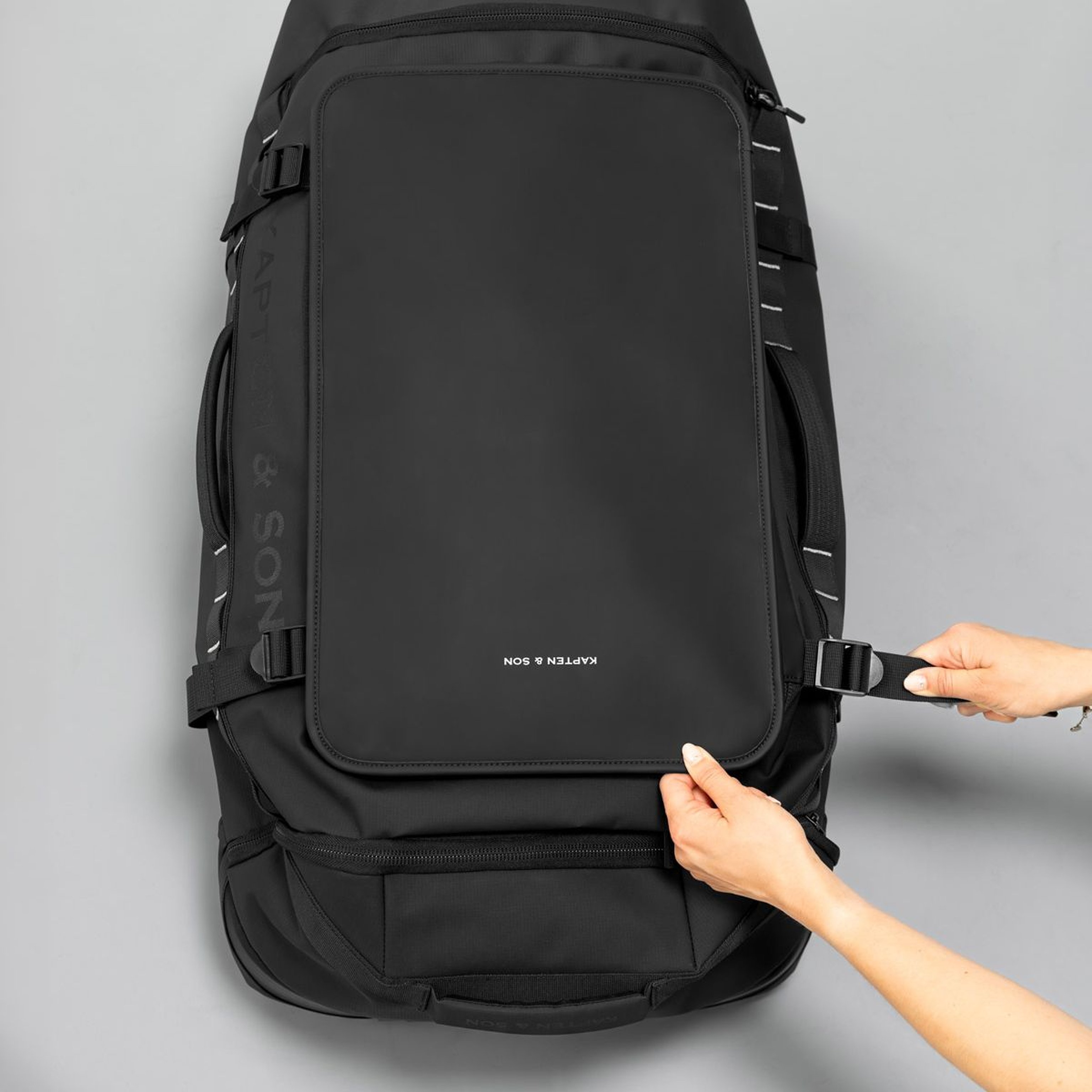 Hands adjusting a buckle on a sleek, black backpack against a gray background. The bag has a minimalist design with visible "KAPTEN & SON" text.