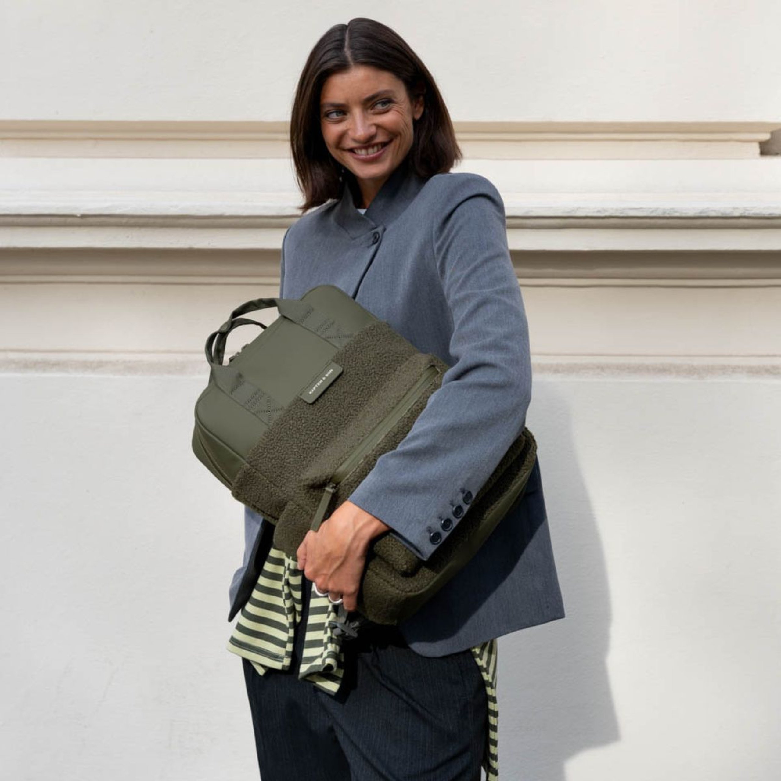 Lächelnde Frau im Blazer hält einen Kapten & Son Rucksack mit weichem Fleece im Arm und steht vor einer Hausfassade.