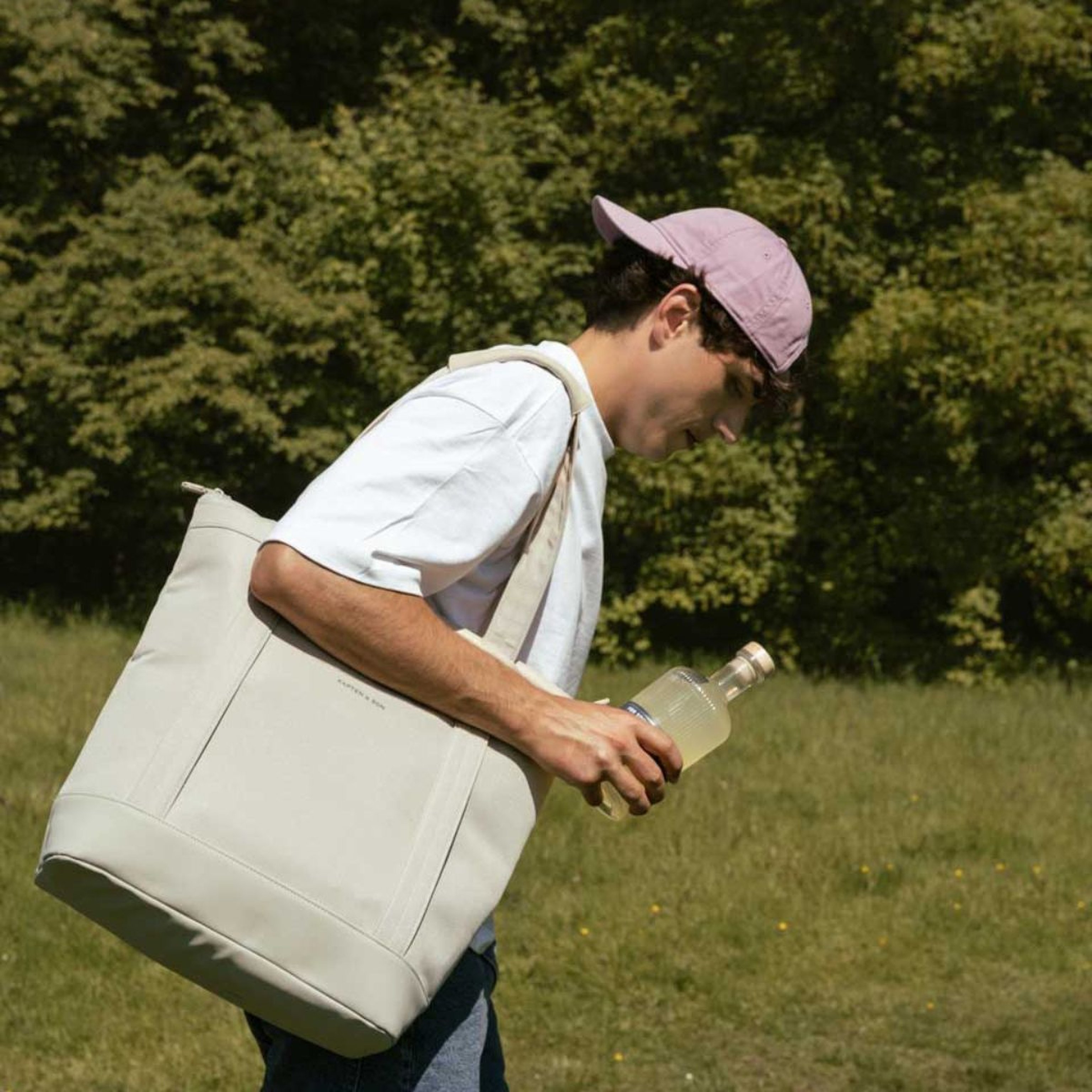 A person in a pink cap and white shirt carries a large beige tote bag and a bottle in a sunny park, with lush green trees in the background.