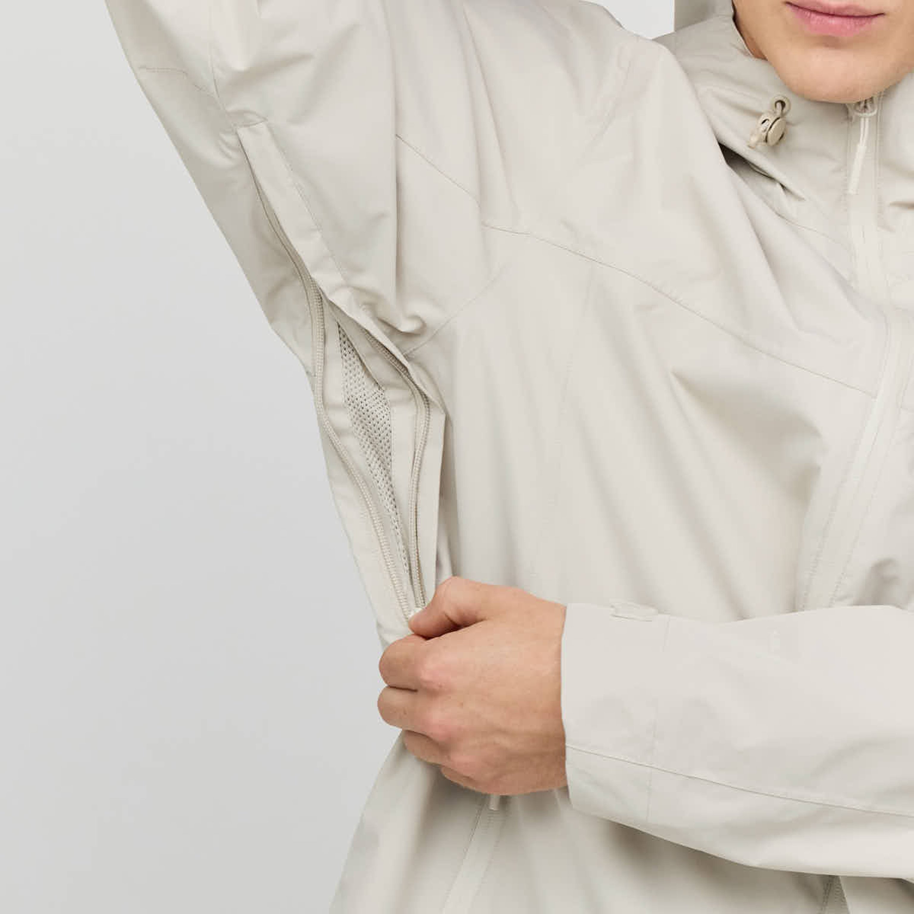 Eine Person in einer hellbeigen Jacke hält diese geöffnet und zeigt eine Netz-Belüftungsöffnung mit Reißverschluss unter dem Arm. Der Fokus liegt auf der Funktionalität und dem Design der Jacke.