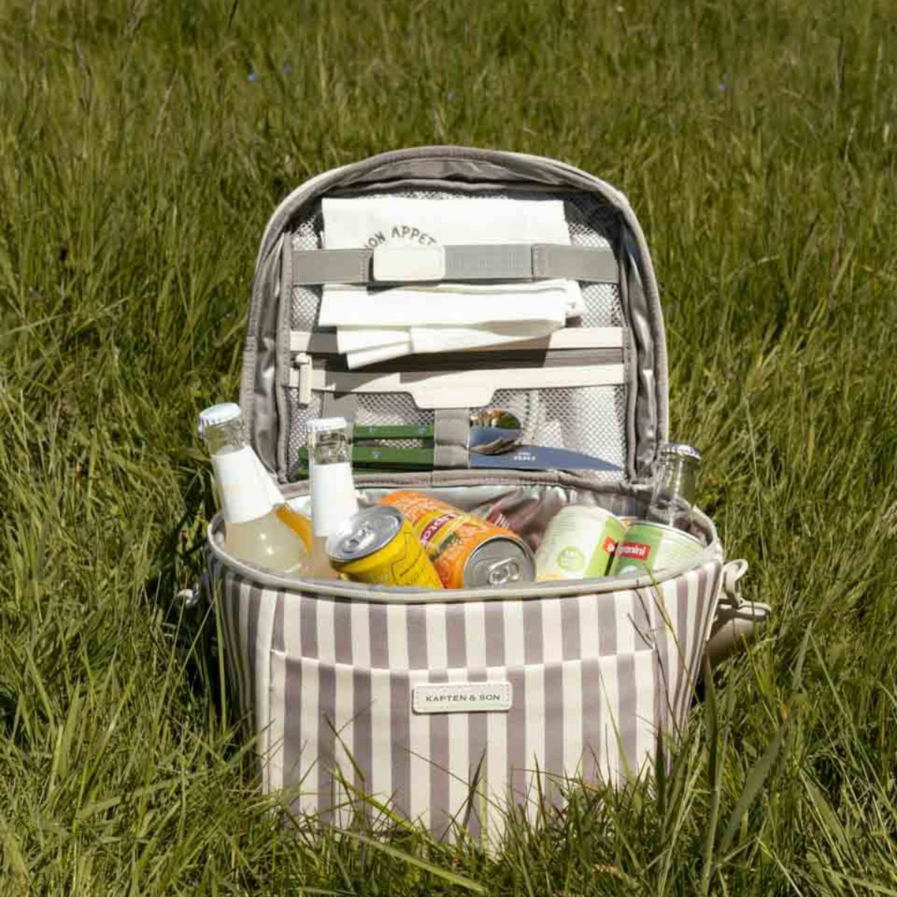 Striped picnic bag open on grass, filled with bottled drinks, soda cans, and cutlery, evoking a cheerful outdoor scene.A person in a white sweater and jeans holds a large tote bag, smiling confidently. The setting is minimalist, conveying a relaxed, stylish tone.