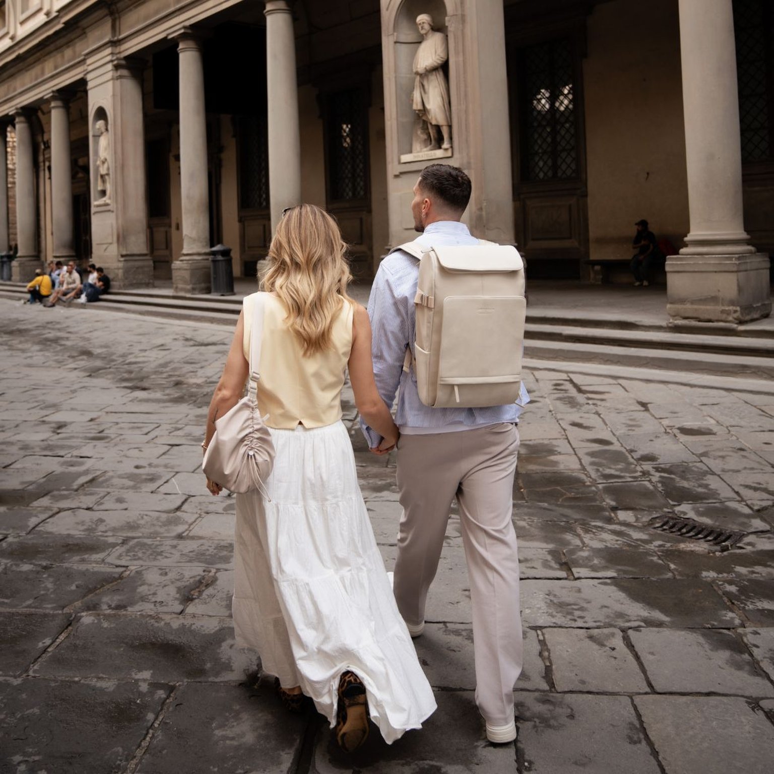 A couple walks hand in hand through a classic European piazza. The man carries a Kapten & Son backpack in Sandstone, paired with a light blue shirt and beige trousers.
