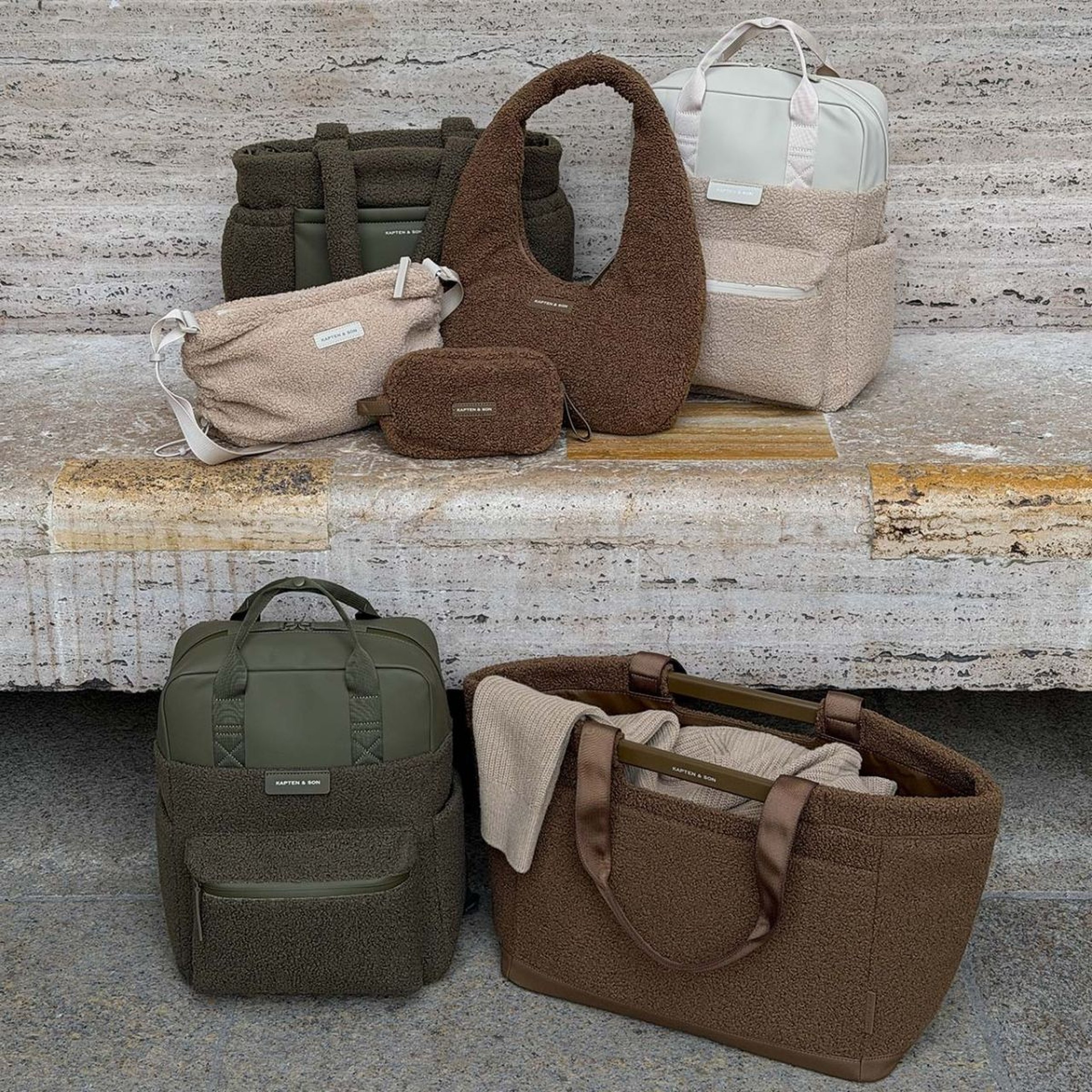 Rucksäcke, Taschen und ein kleines Täschchen von Kapten & Son mit flauschiger Teddy-Oberfläche, auf und vor einer Steinbank arrangiert.