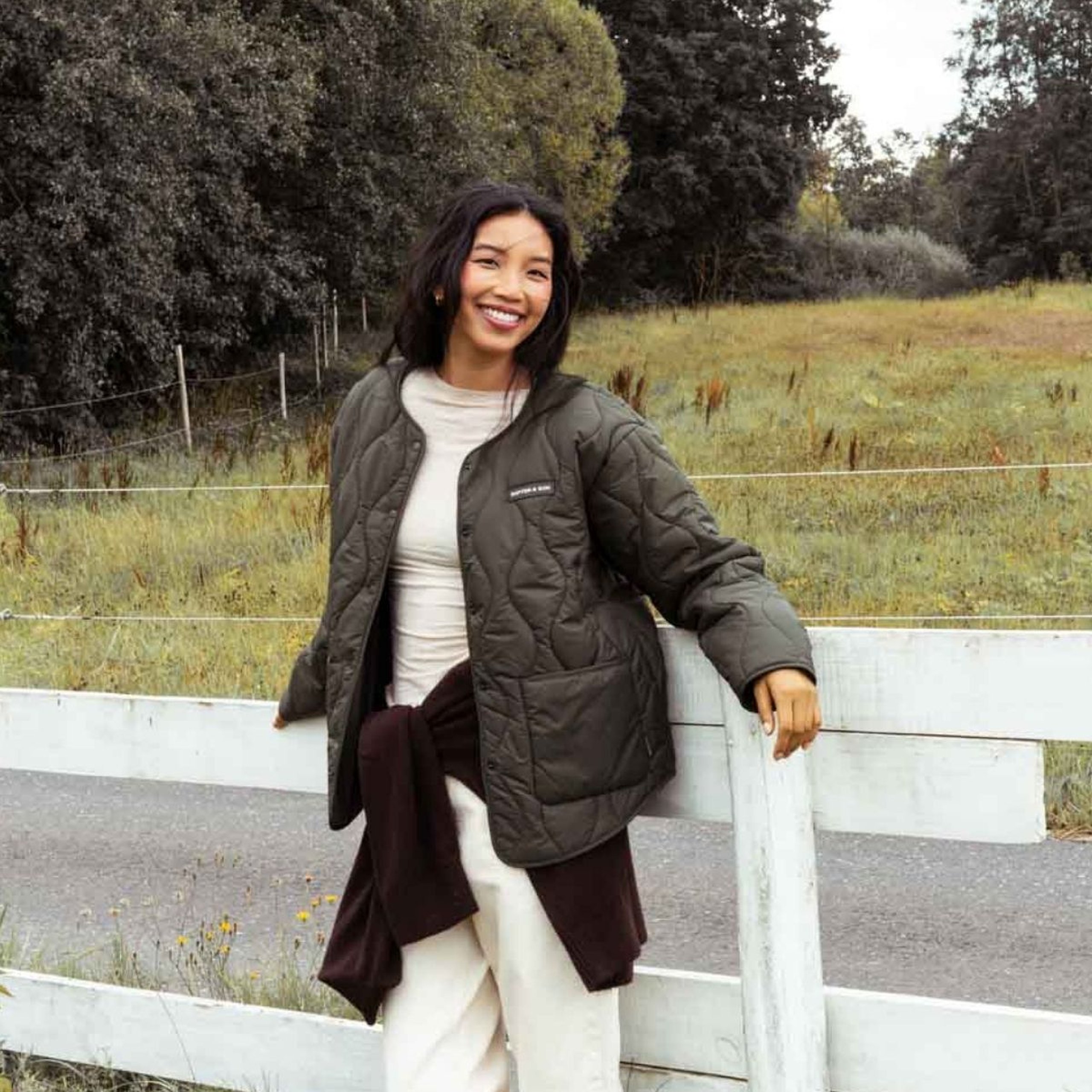 Woman wearing a quilted jacket leaning against a wooden fence in a rural outdoor setting.