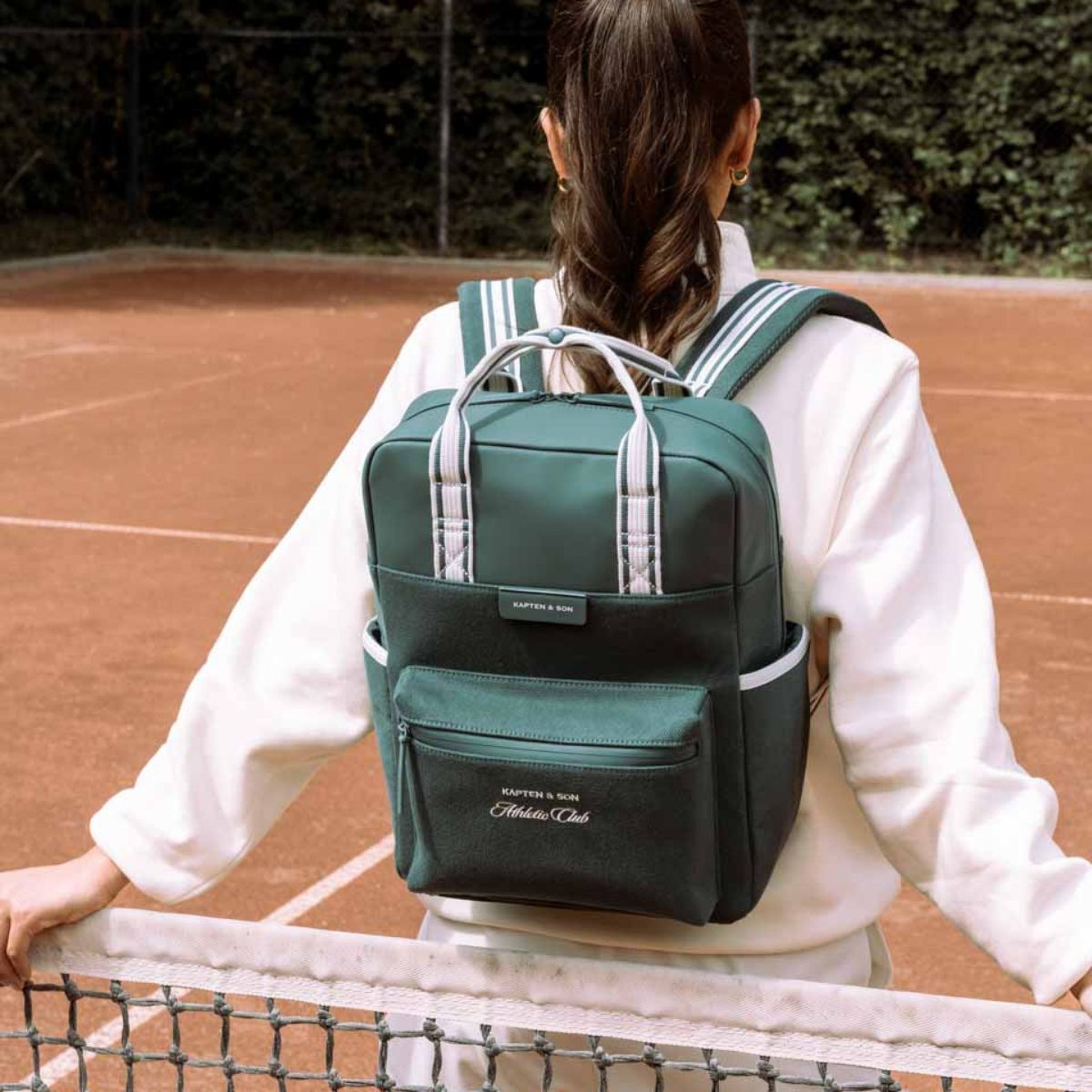 Eine Person mit Pferdeschwanz trägt ein weißes Outfit und einen grünen Rucksack und steht an einem Tennisnetz. Bäume bilden einen üppigen Hintergrund unter einem klaren Himmel.