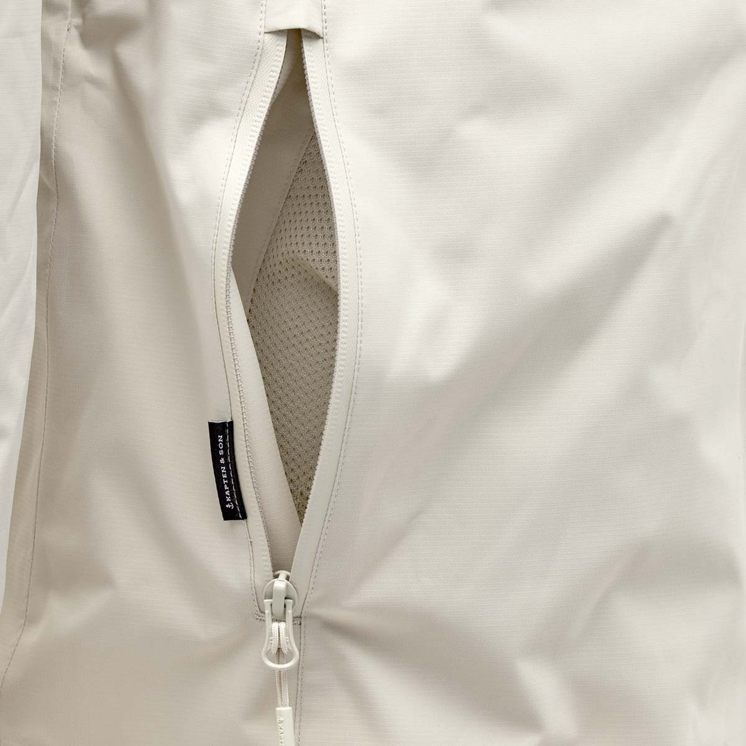 Nahaufnahme einer beigefarbenen Jacke mit offenem Reißverschluss, die ein Netzfutter zeigt. Die Textur ist glatt, und ein schwarzes Stoffetikett ist in der Nähe des Reißverschlusses angebracht.