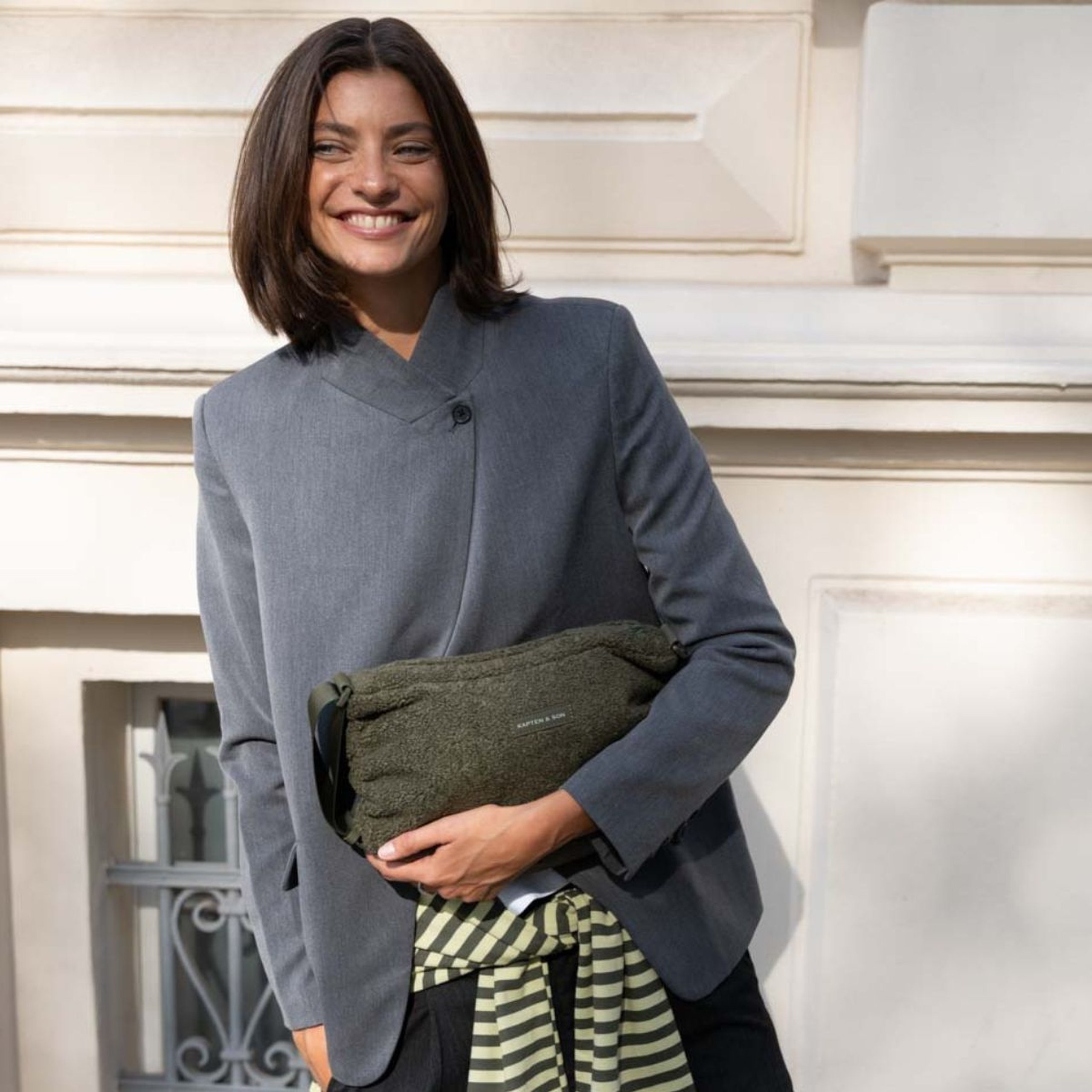 Lächelnde Frau im Blazer hält eine Kapten & Son Tasche aus weichem Fleece unter dem Arm und steht vor einer Hausfassade.