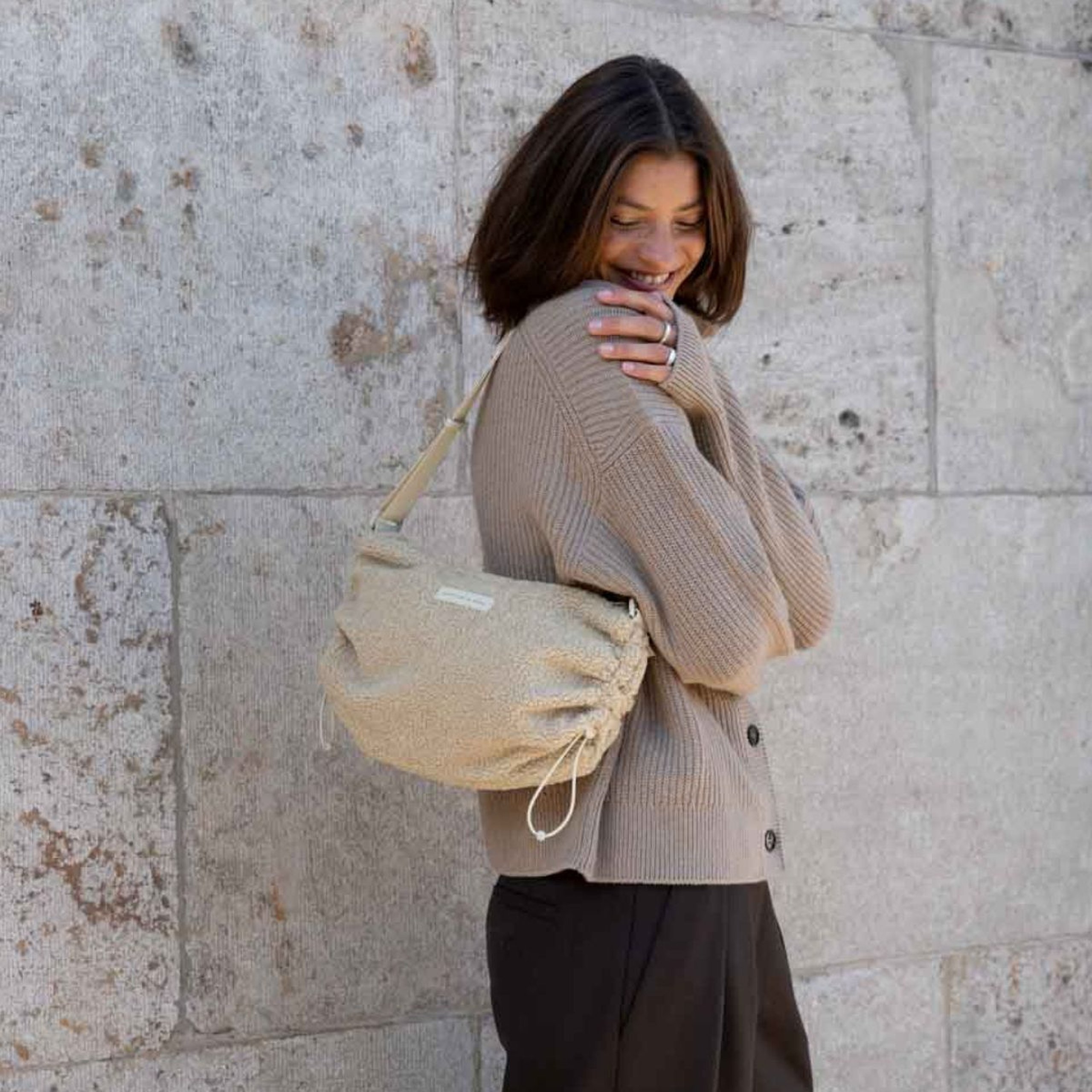Lächelnde Frau in einer Strickjacke trägt eine Kapten & Son Schultertasche aus weichem Fleece mit seitlichem Kordelzug vor einer Steinwand.