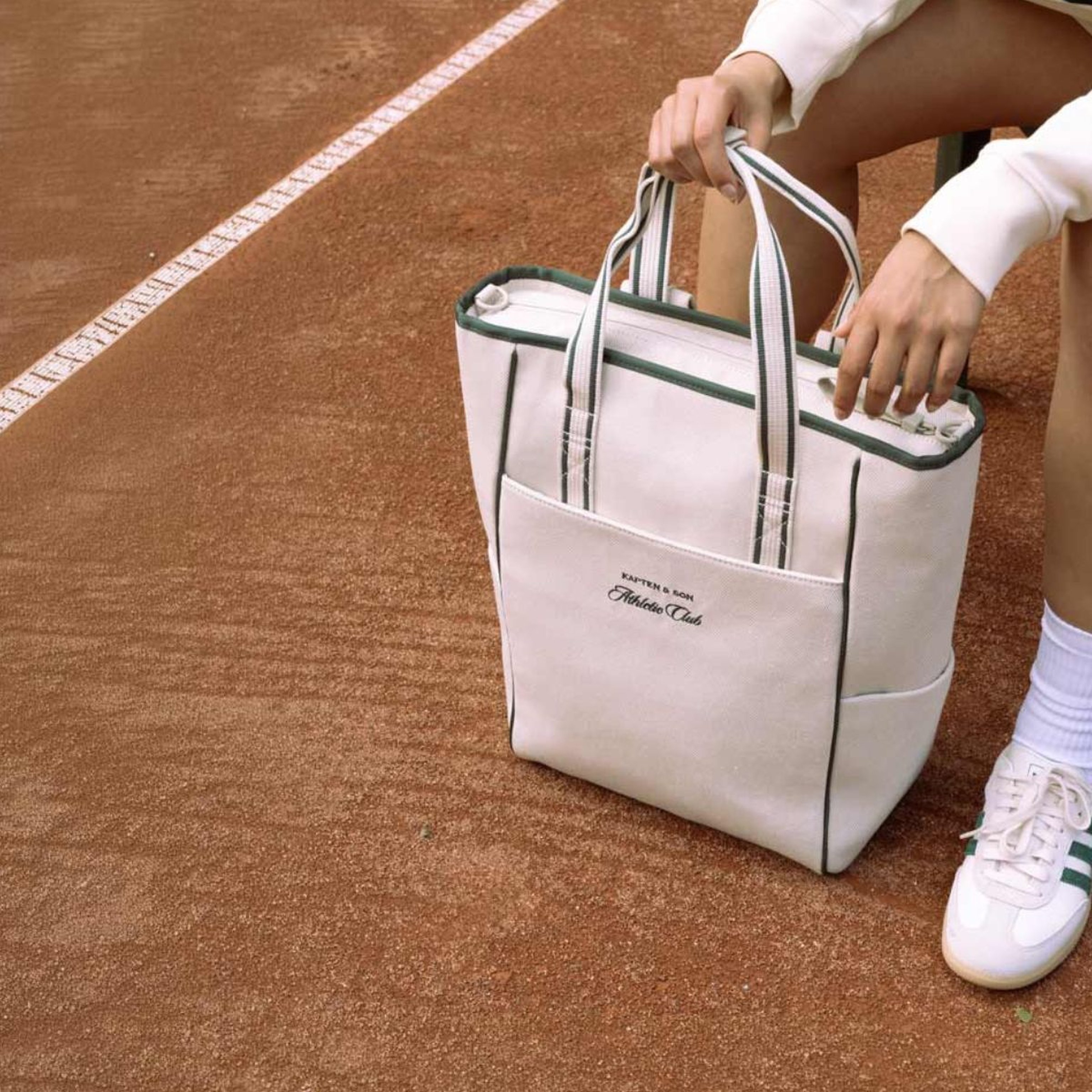 Eine Person in weißem Pullover und Sneakers sitzt auf einem Tennisplatz und hält eine stilvolle weiße Tote Bag mit grünen Details – eine sportlich-entspannte Atmosphäre.