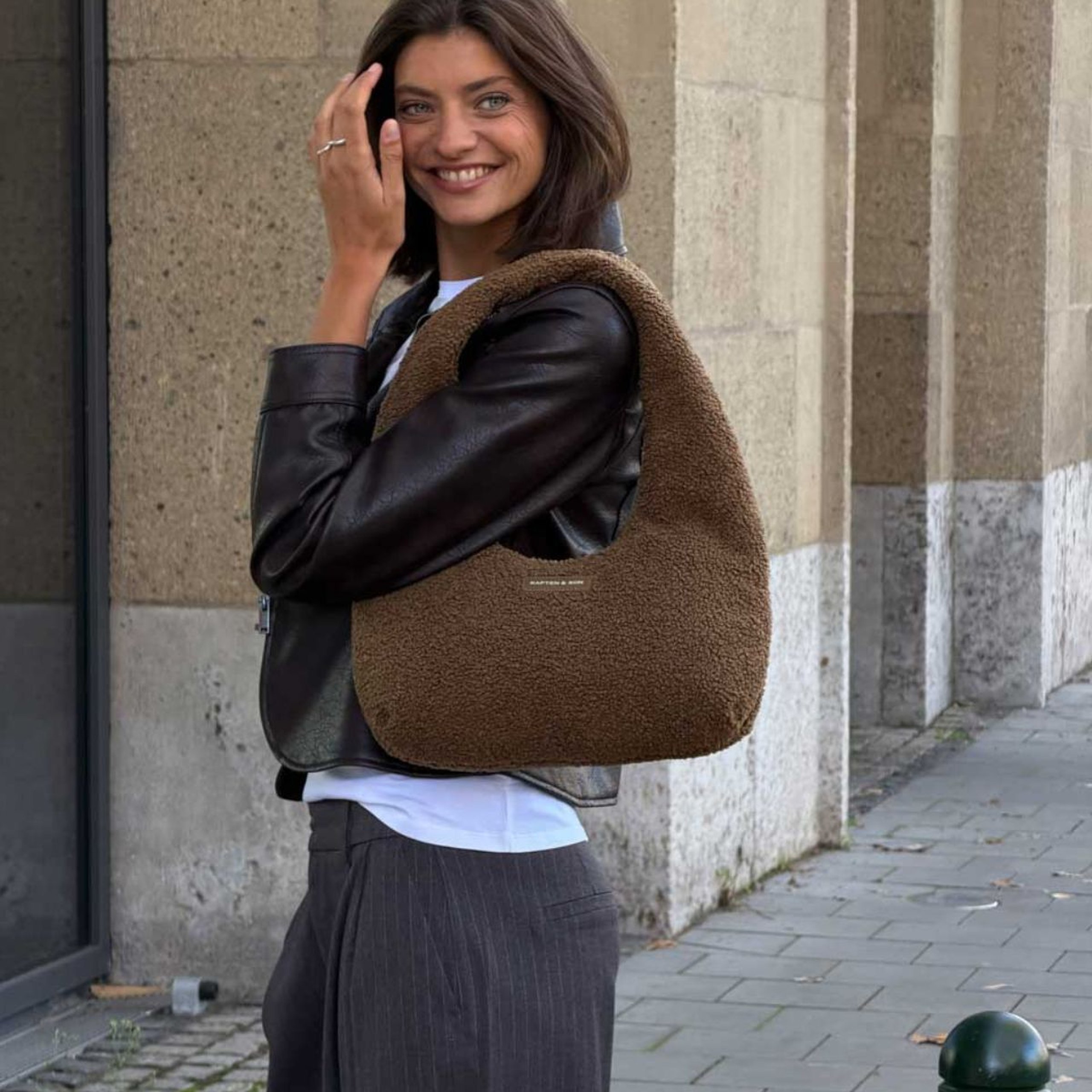 Lächelnde Frau in Lederjacke trägt eine große Kapten & Son Schultertasche aus weichem Fleece vor einer Steinfassade.