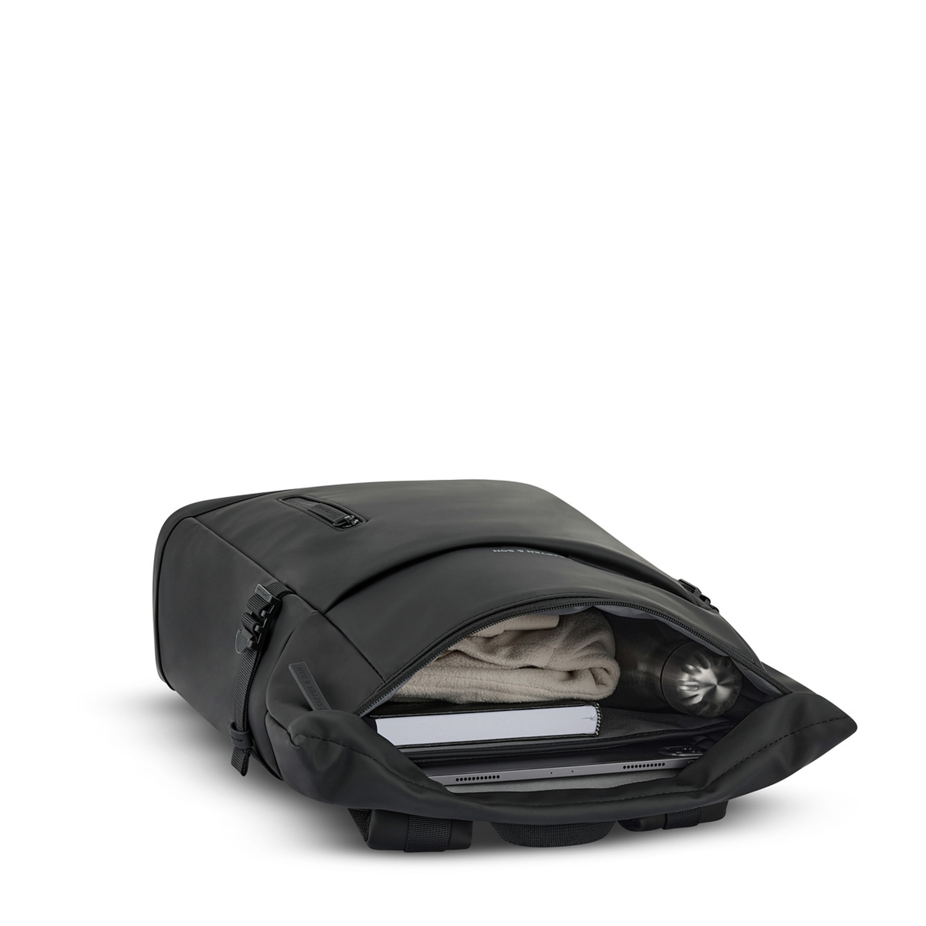 Ein schlichter schwarzer Rucksack liegt geöffnet da und gibt den Blick auf eine ordentlich gefaltete beige Jacke, eine silberne Wasserflasche, ein Notizbuch und elektronische Geräte frei – auf weißem Hintergrund.