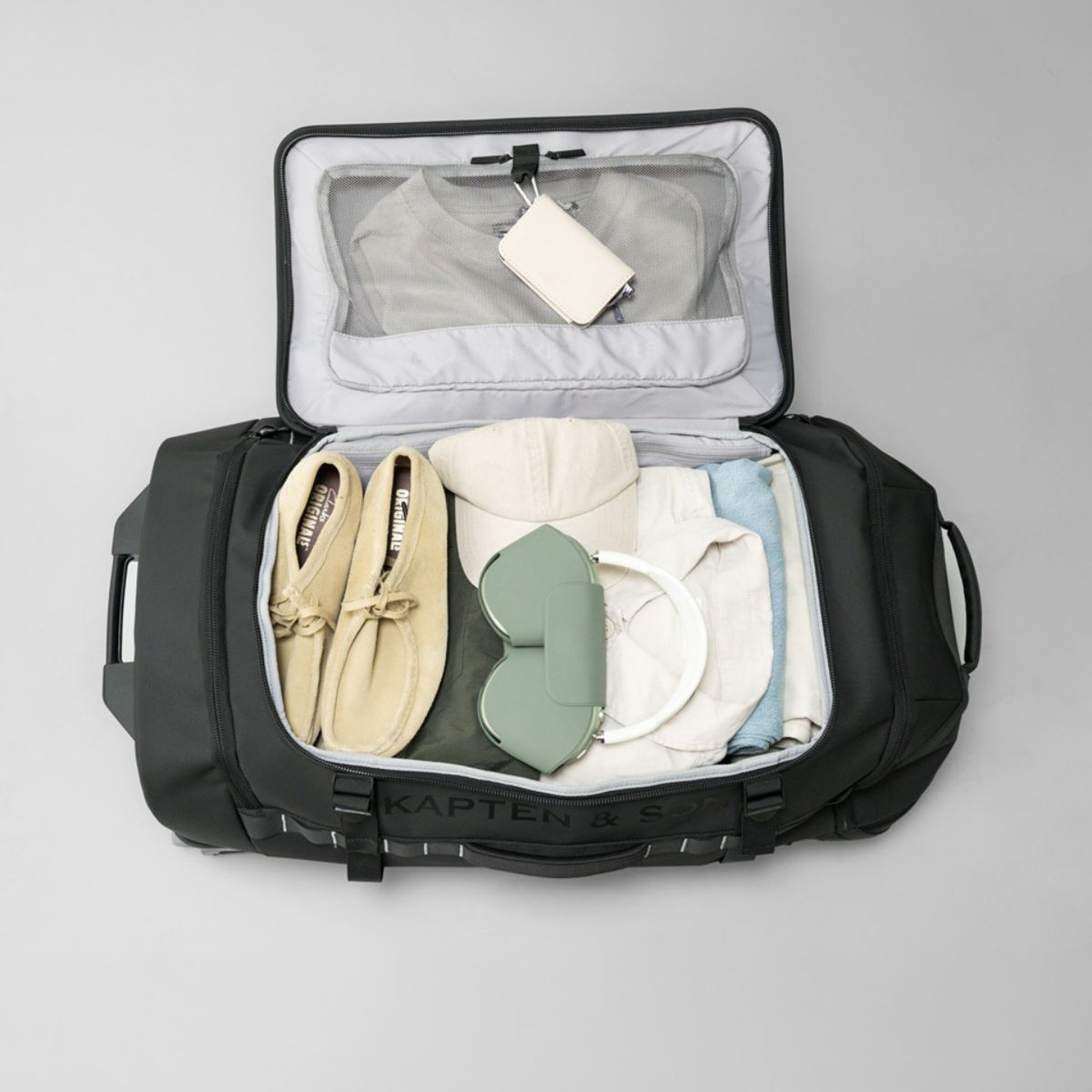 Open black duffel bag with organized contents: beige shoes, folded clothes, a cap, and green headphones. A light, travel-ready vibe.
