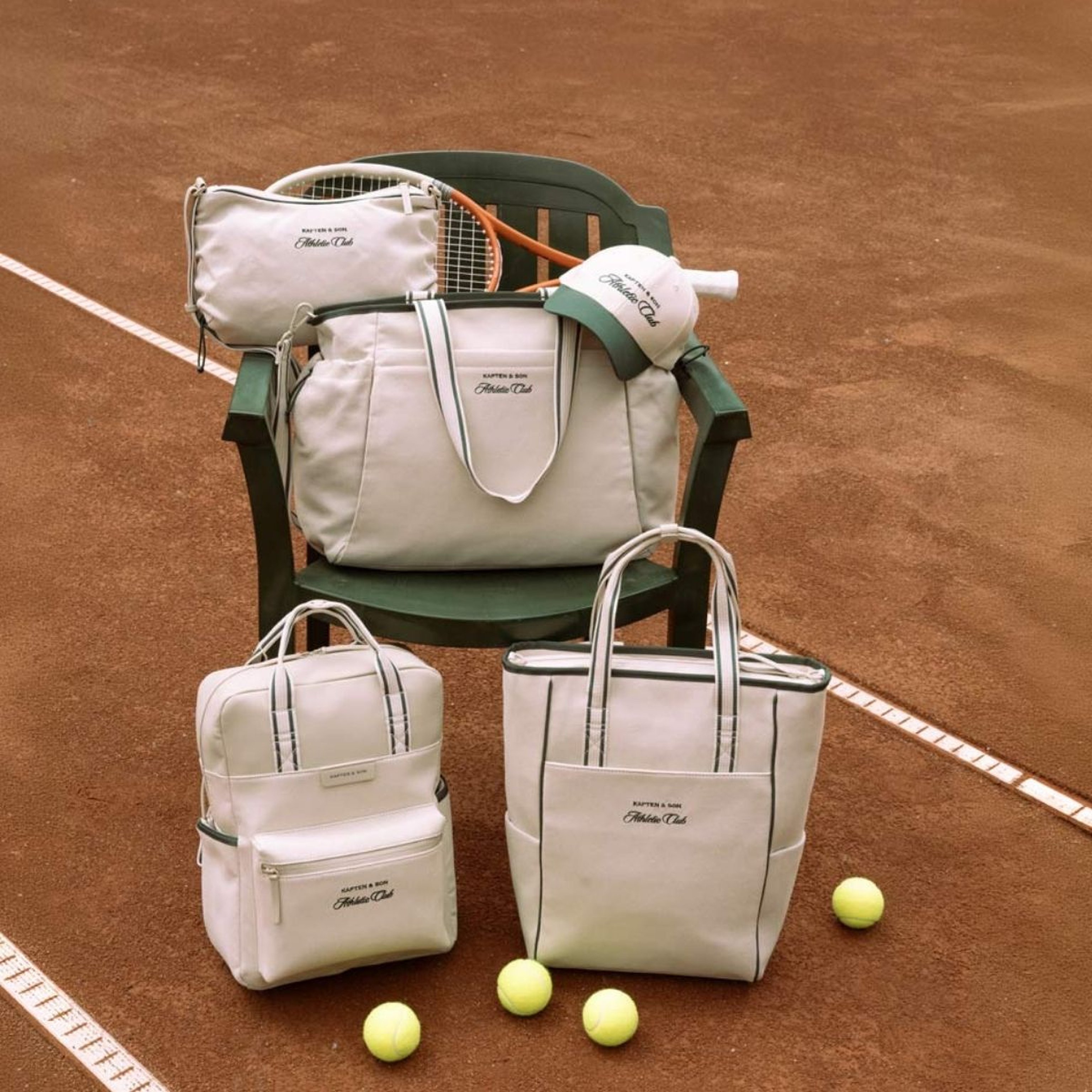 Grüner Stuhl auf einem Sandplatz mit weißen Tennistaschen, einer Cap und einem Schläger. Drei Tennisbälle liegen auf dem Boden und vermitteln eine sportliche Eleganz.