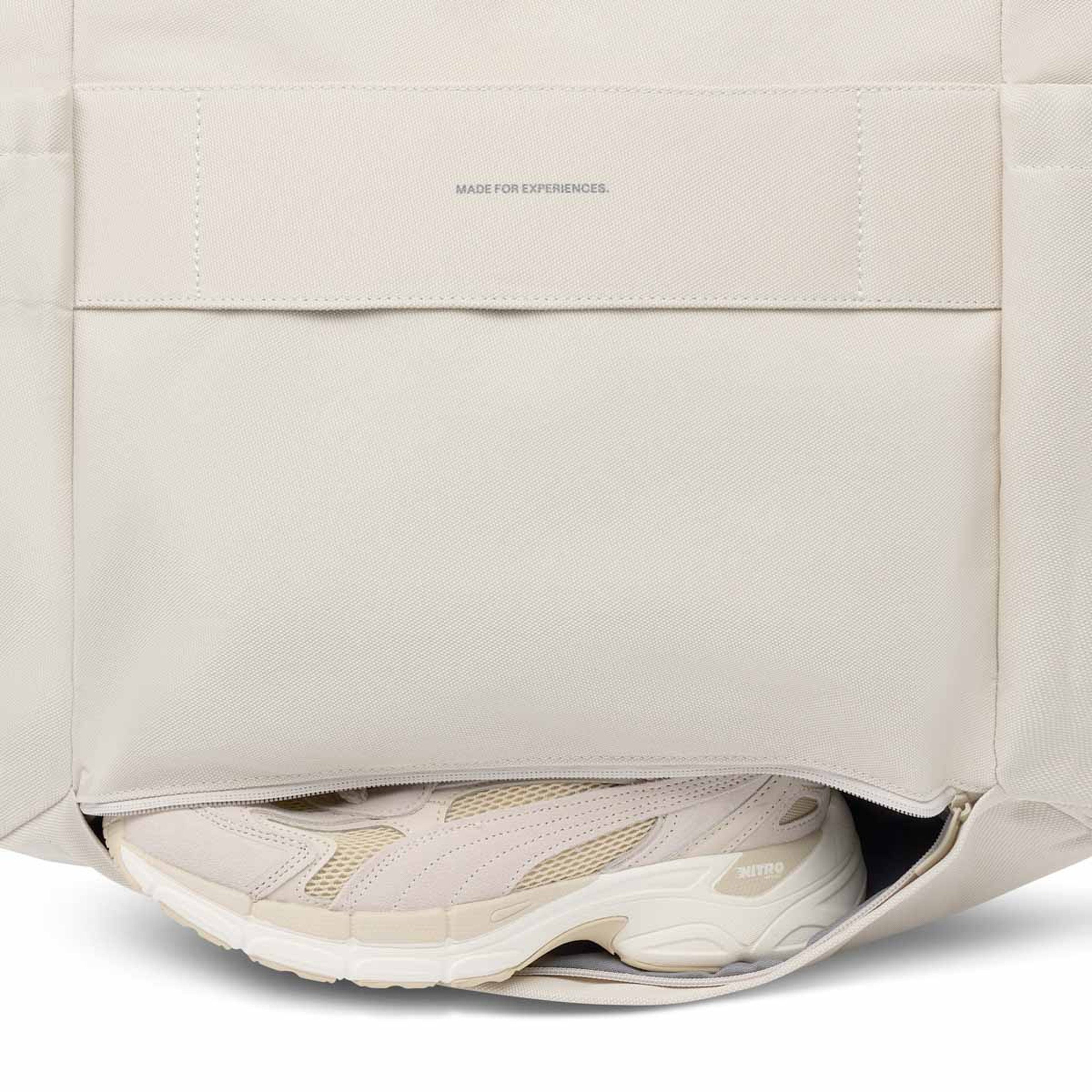 Unteres Reißverschlussfach einer sandfarbenen Tasche mit einem Paar beigefarbener Sportschuhe darin.