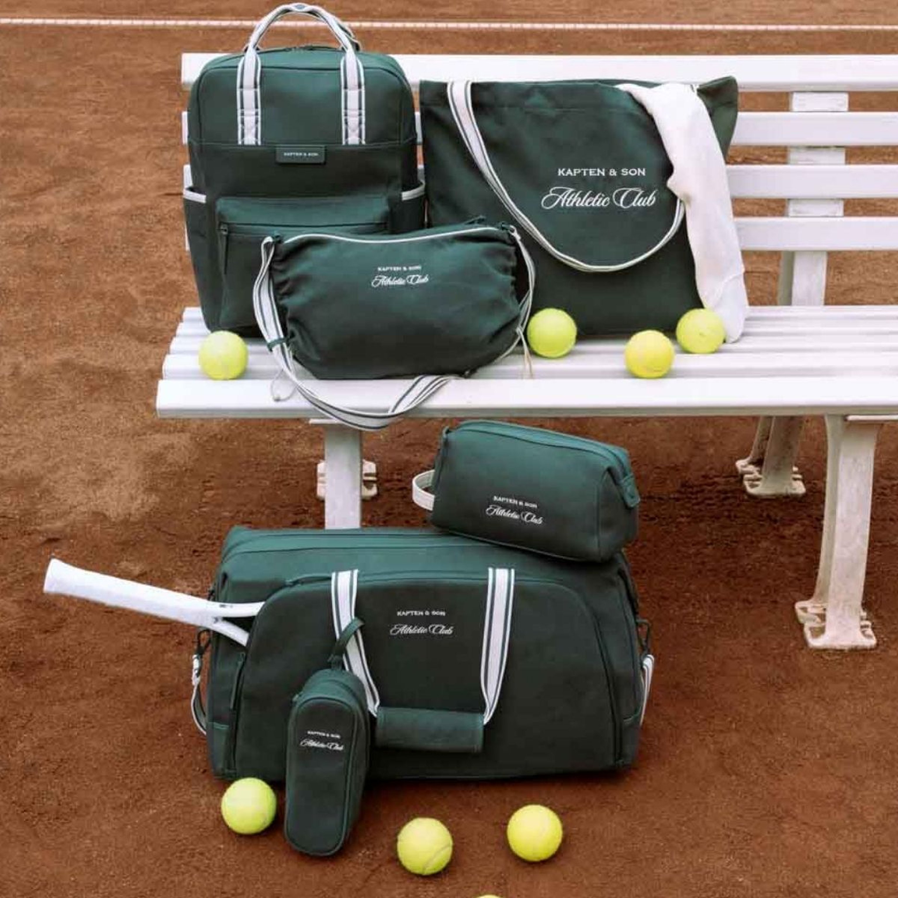 Grüne Sporttaschen und Tennisbälle sind auf einer weißen Bank auf einem Sandplatz arrangiert. Die Szene vermittelt eine organisierte, sportliche Atmosphäre.