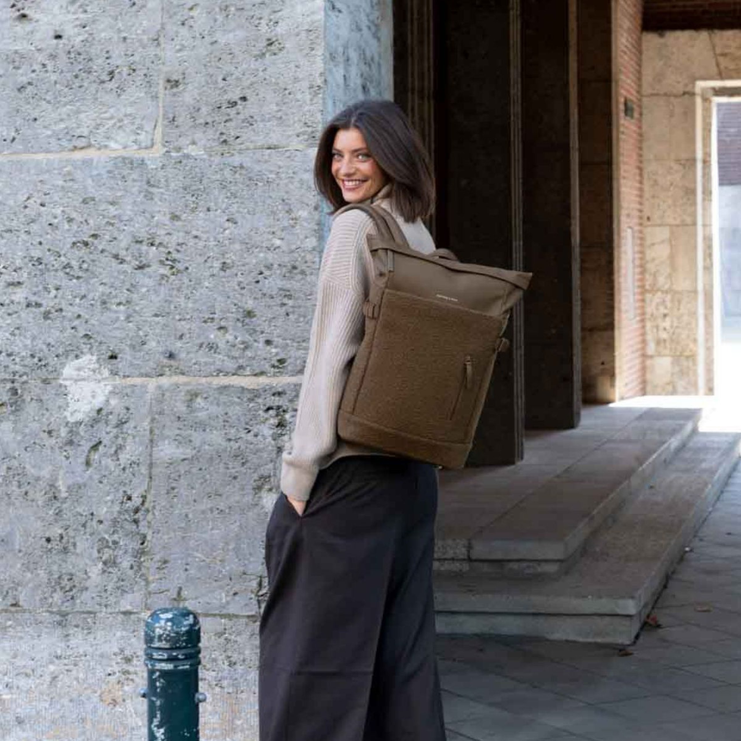 Lächelnde Frau in einer Strickjacke trägt einen Kapten & Son Rolltop-Rucksack mit weichem Fleece und blickt über die Schulter zurück.
