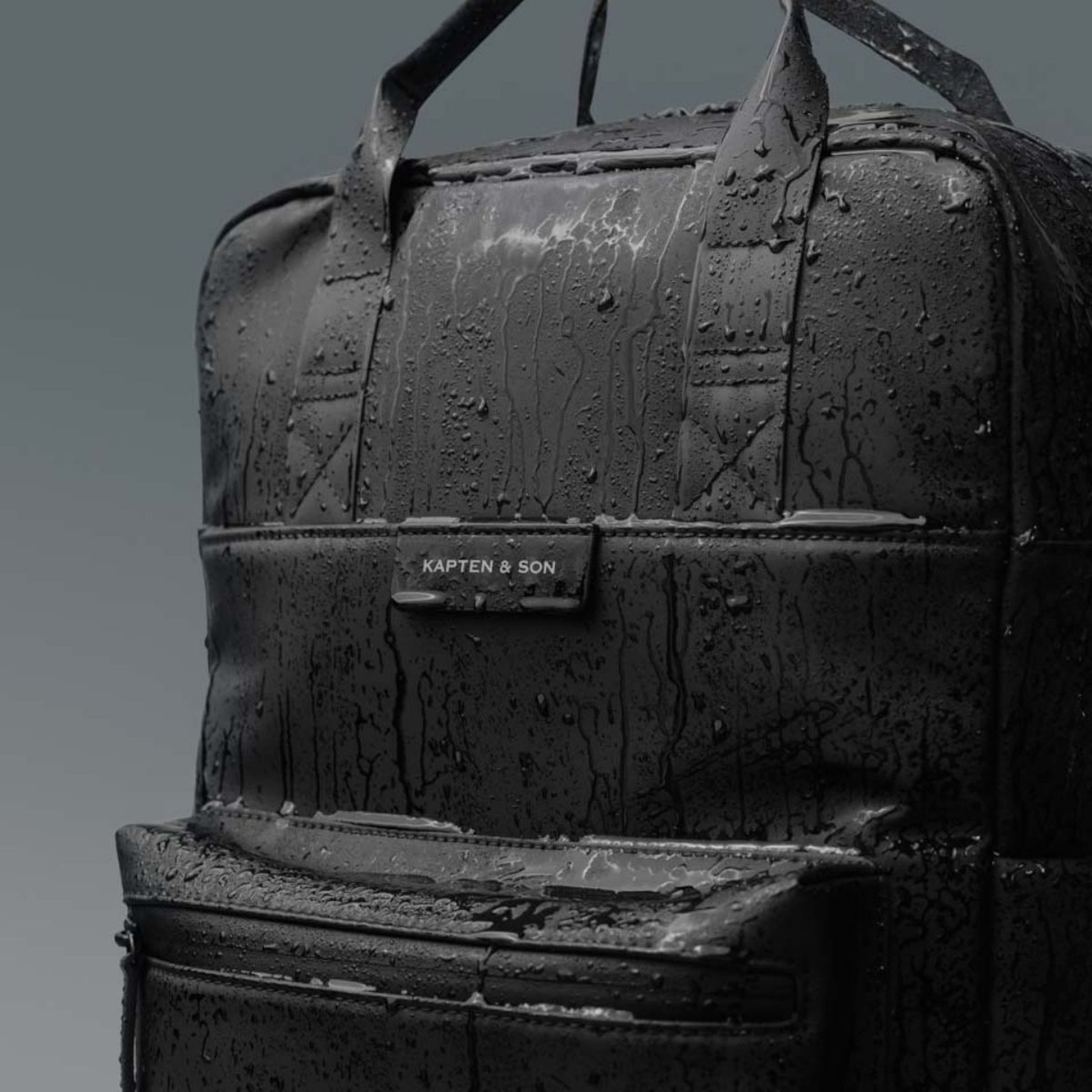 Close-up of a black Kapten & Son backpack covered in raindrops, highlighting its water-resistant material. The scene conveys a sleek, durable vibe.