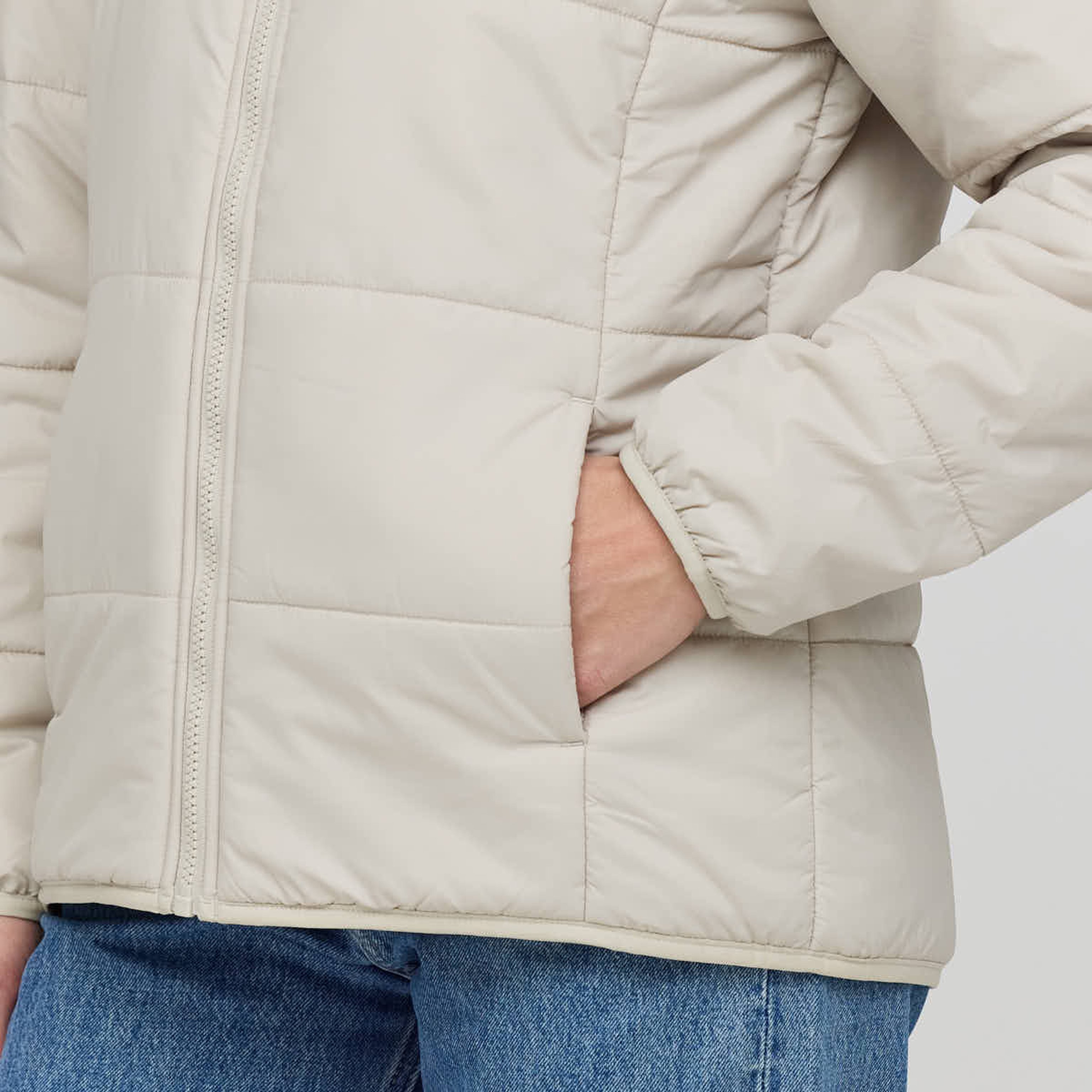 Person wearing a cream-colored, quilted jacket with a visible zipper and hand in pocket, paired with blue jeans. The tone is casual and cozy.