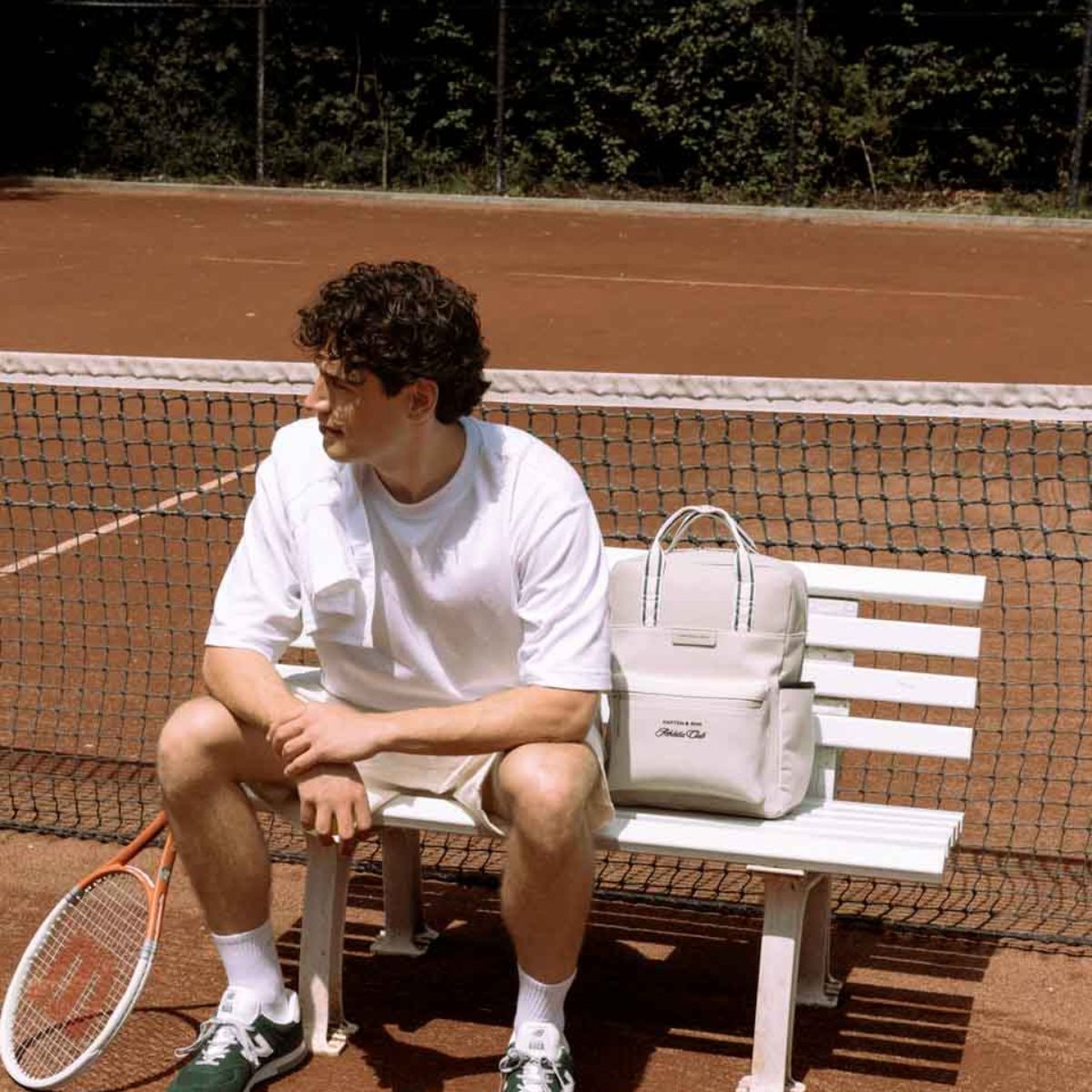 Zwei Personen stehen am Tennisnetz und lächeln. Eine hält einen Schläger, trägt einen grünen Pullover und Shorts. Die andere trägt einen Rucksack und ein weißes Outfit.Ein Mann in weißem T-Shirt und Shorts sitzt auf einer Bank an einem Tennisplatz und hält einen Schläger. Ein beiger Rucksack liegt neben ihm. Die Szene wirkt entspannt.