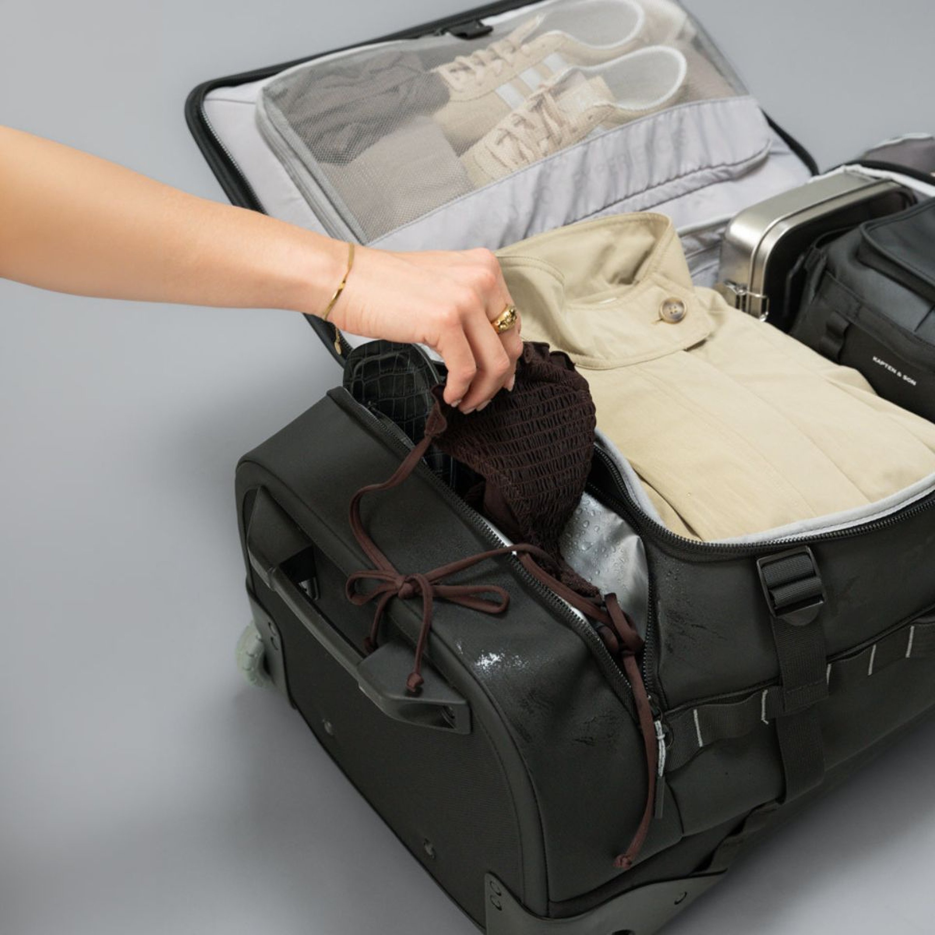 A person places folded clothes and shoes inside a black suitcase on wheels. The scene suggests organized packing, with a neat and tidy arrangement.