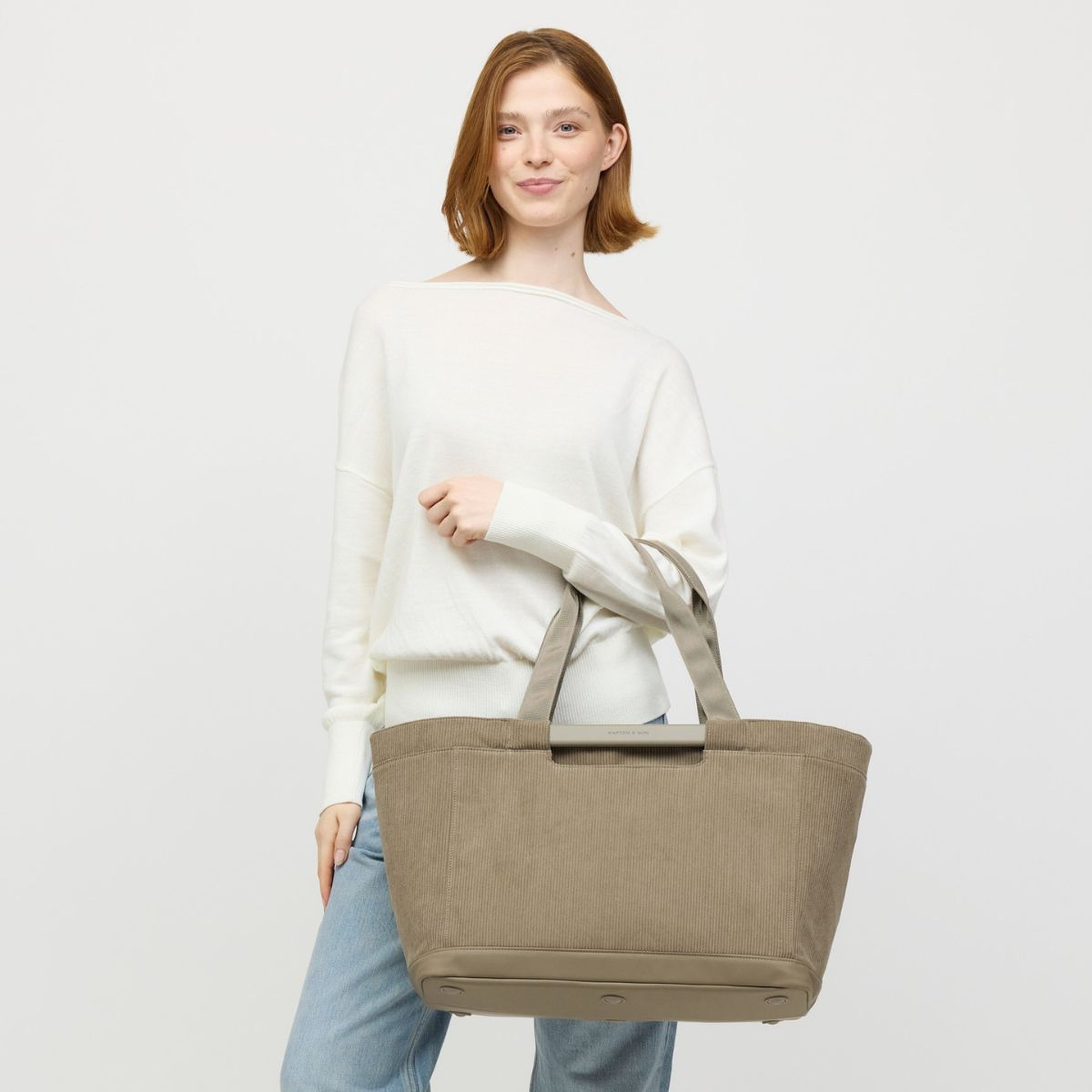 A person in a white sweater and jeans holds a large tote bag, smiling confidently. The setting is minimalist, conveying a relaxed, stylish tone.