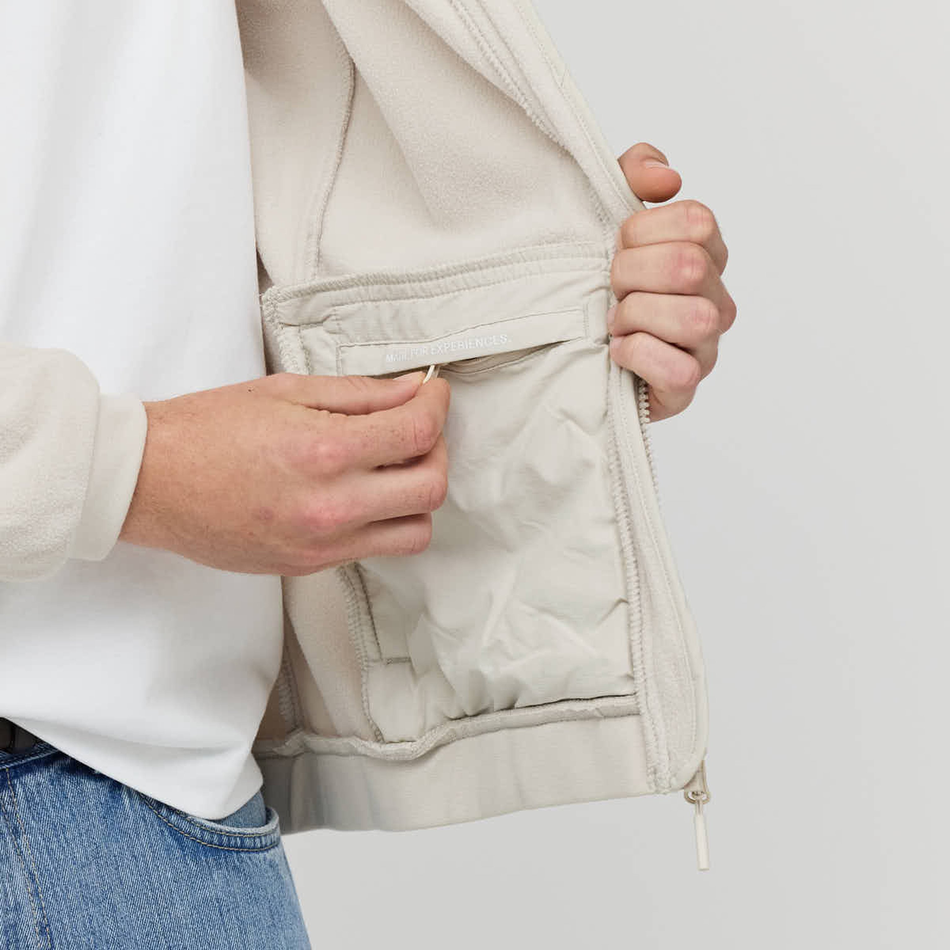 A person in a cream jacket demonstrates opening a small interior zipper pocket. The focus is on the hands and pocket, highlighting practicality and design.