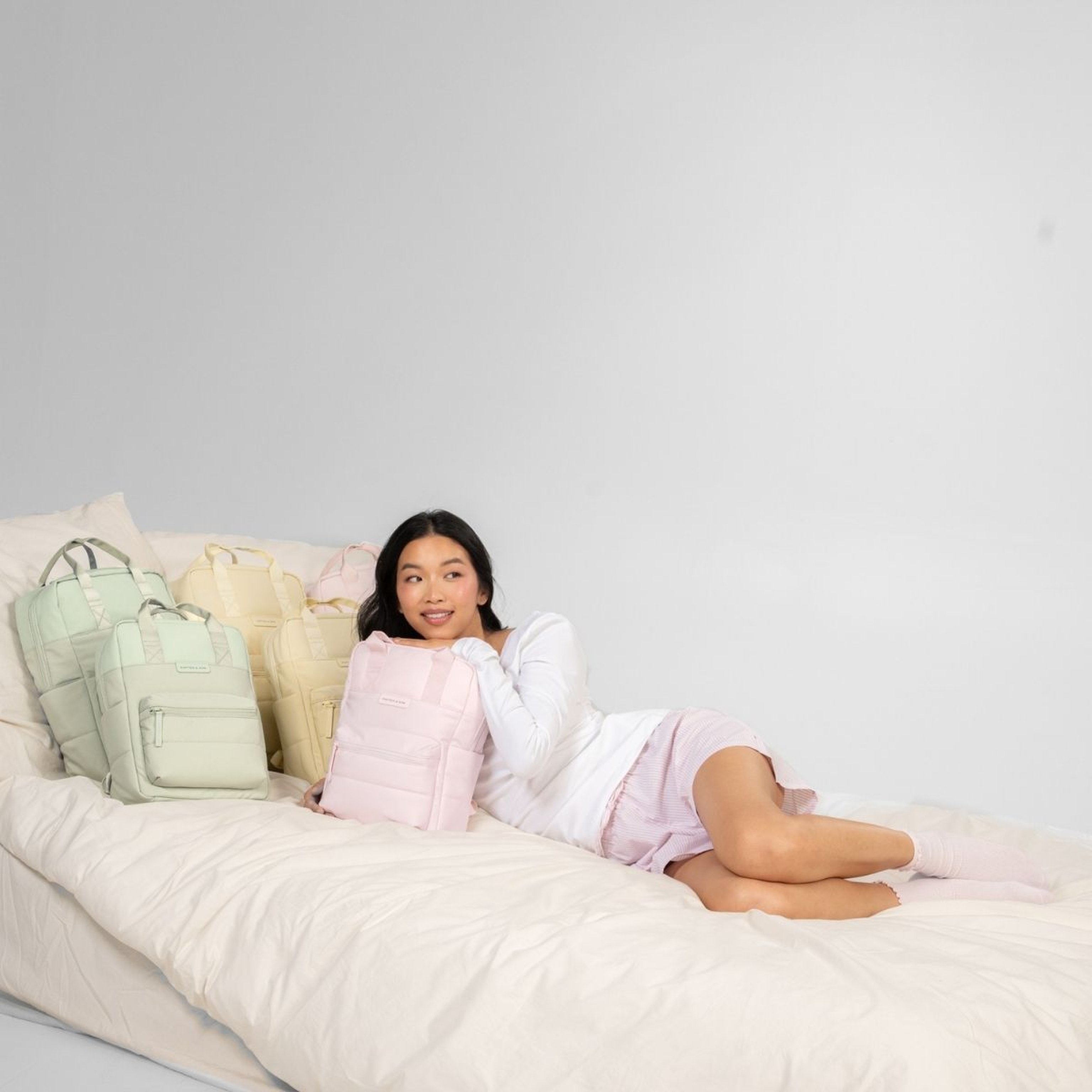 Eine Frau kuschelt sich unter eine weiße Bettdecke und lächelt mit einem pastellrosa Rucksack. Hinter ihr sind sechs weitere Rucksäcke in zartem Rosa, Gelb und Grün arrangiert.