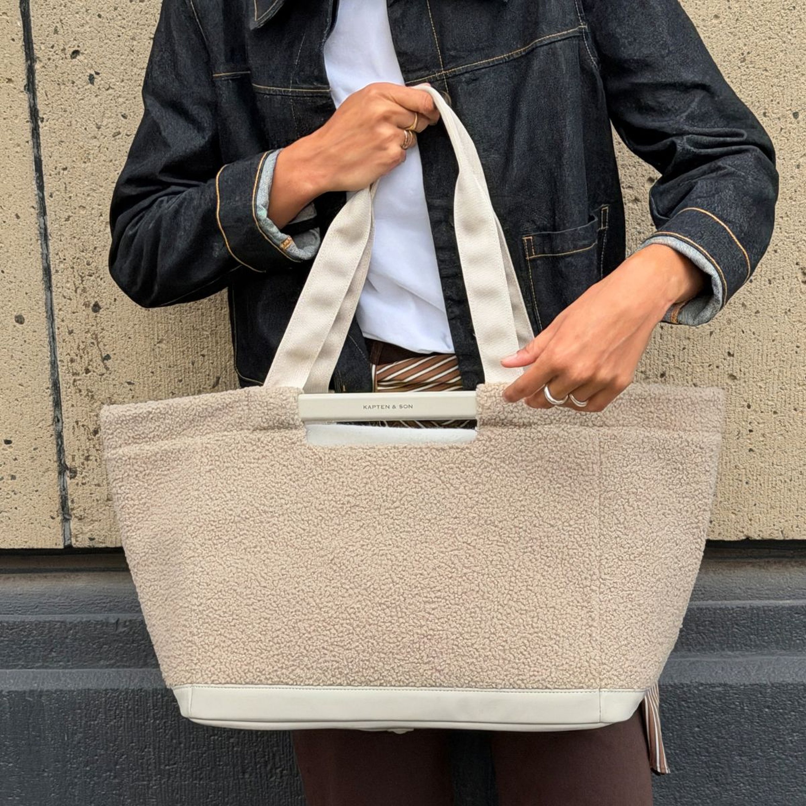 Nahaufnahme einer Person in Jeansjacke, die eine große Kapten & Son Tasche aus weichem Fleece an den Tragehenkeln hält.
