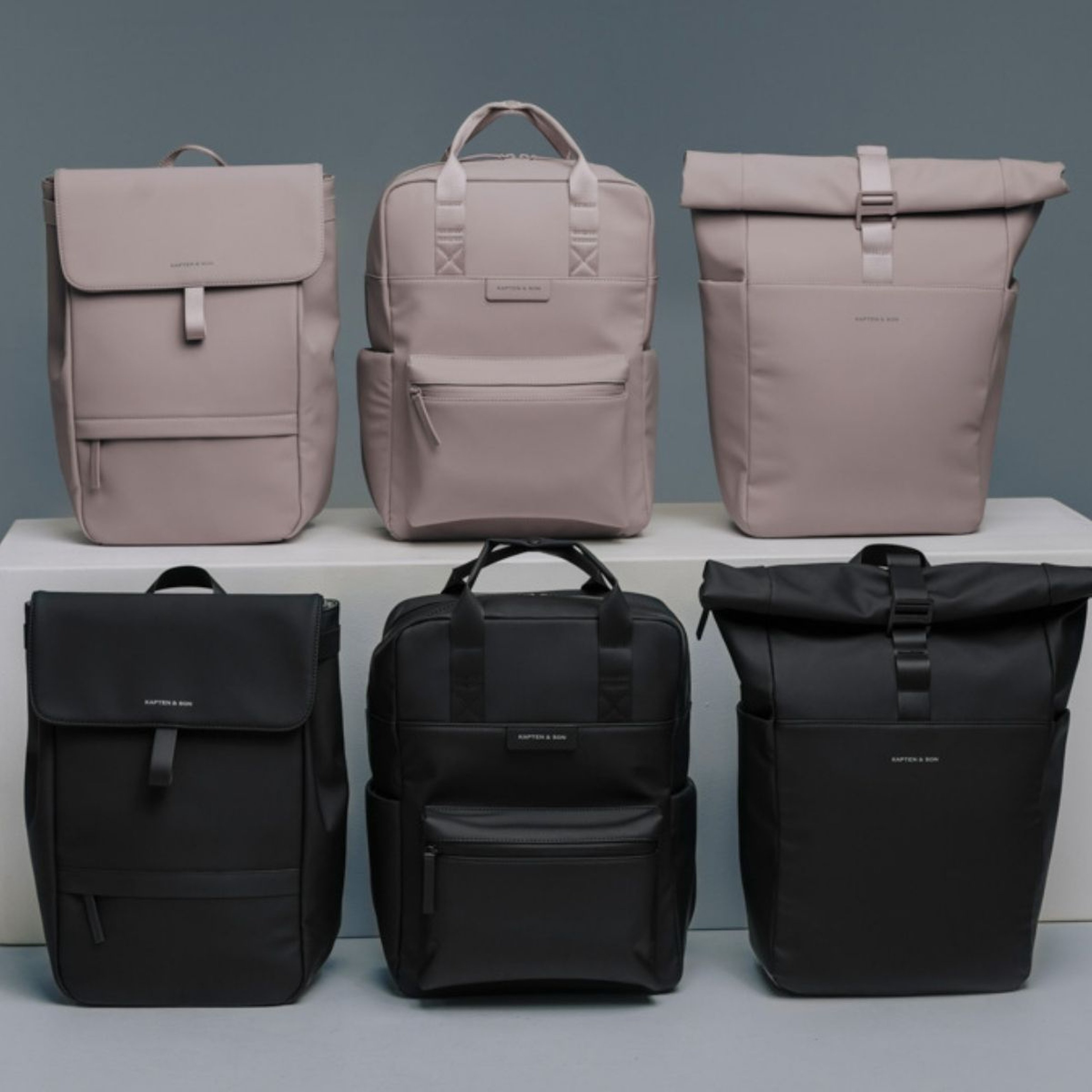 A collection of six minimalist backpacks in two rows against a gray backdrop. The top row features three light beige bags; the bottom row showcases three black bags.