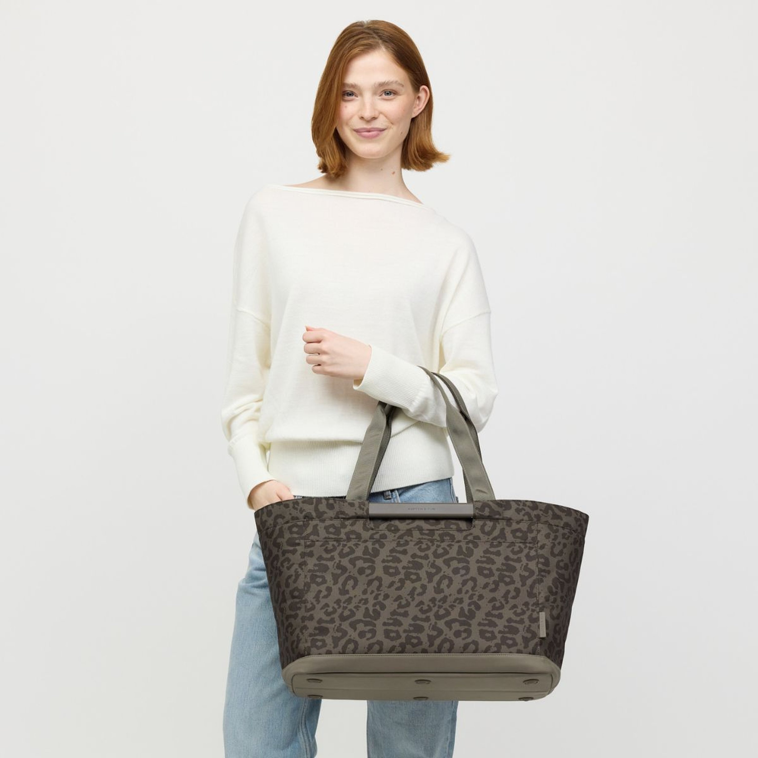 A person in a white sweater and jeans holds a large tote bag, smiling confidently. The setting is minimalist, conveying a relaxed, stylish tone.
