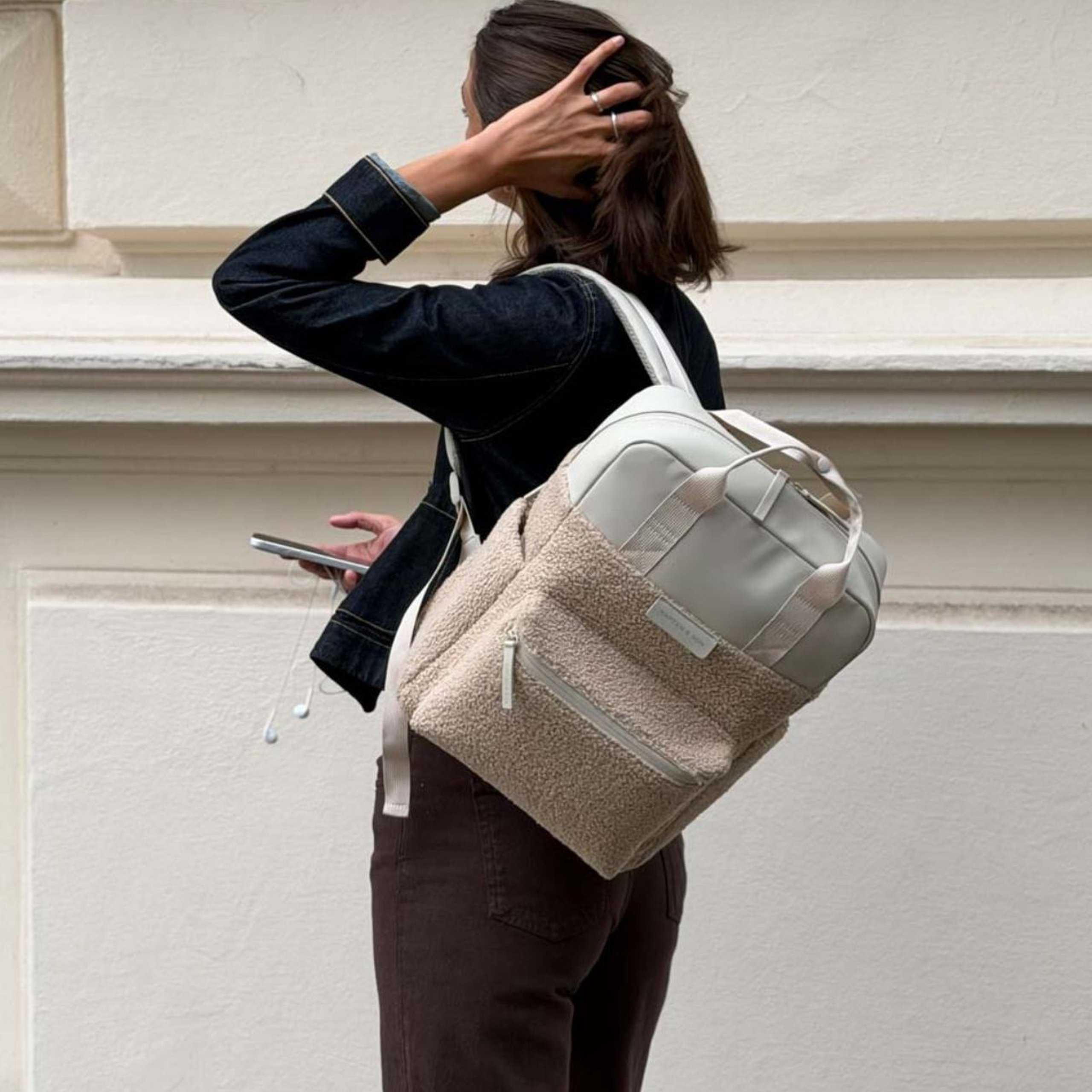 Frau in Jeansjacke trägt einen Kapten & Son Rucksack mit weichem Fleece über einer Schulter und hält ein Smartphone in der Hand.