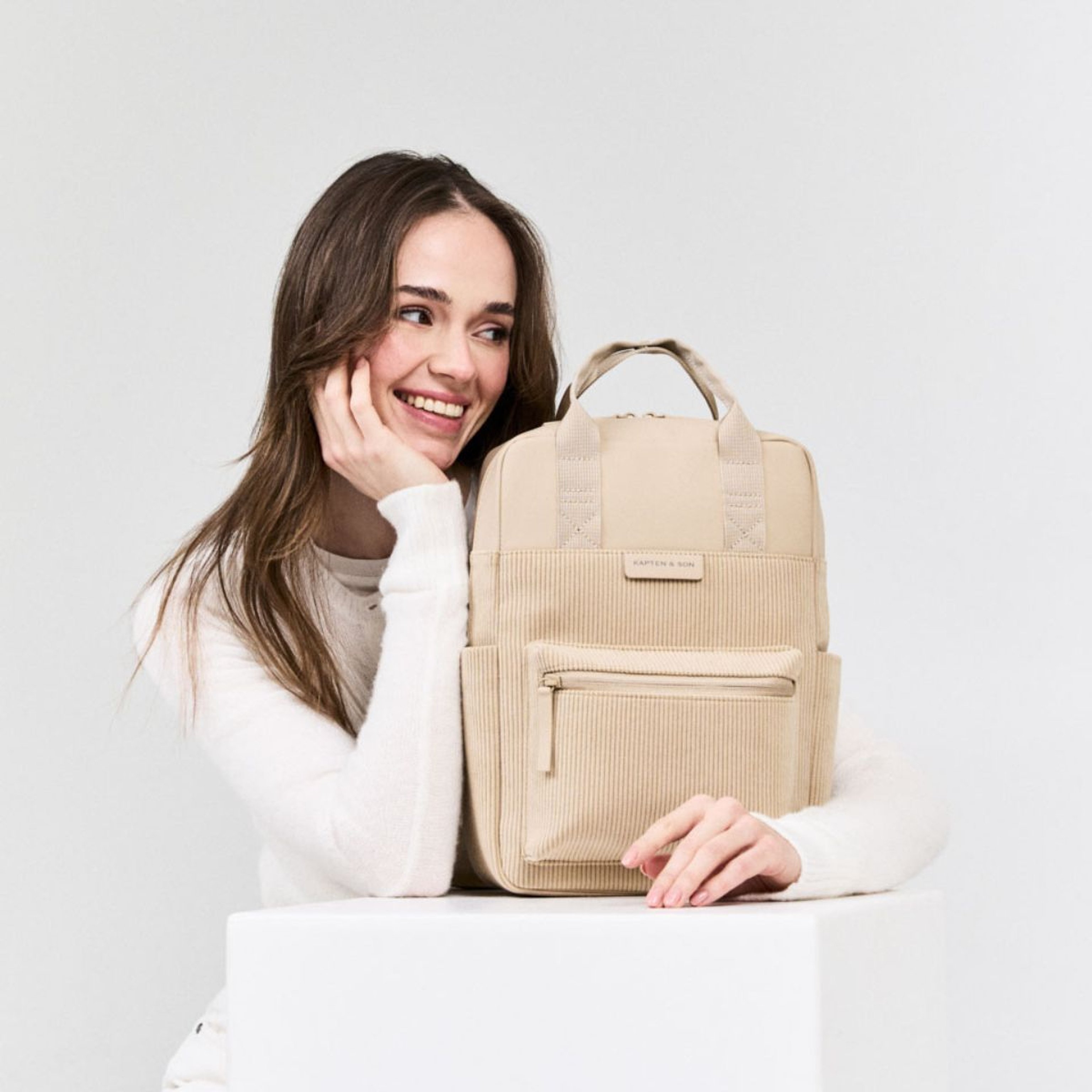 Una donna sorridente tiene in mano uno zaino beige, trasmettendo un’atmosfera allegra e casual.