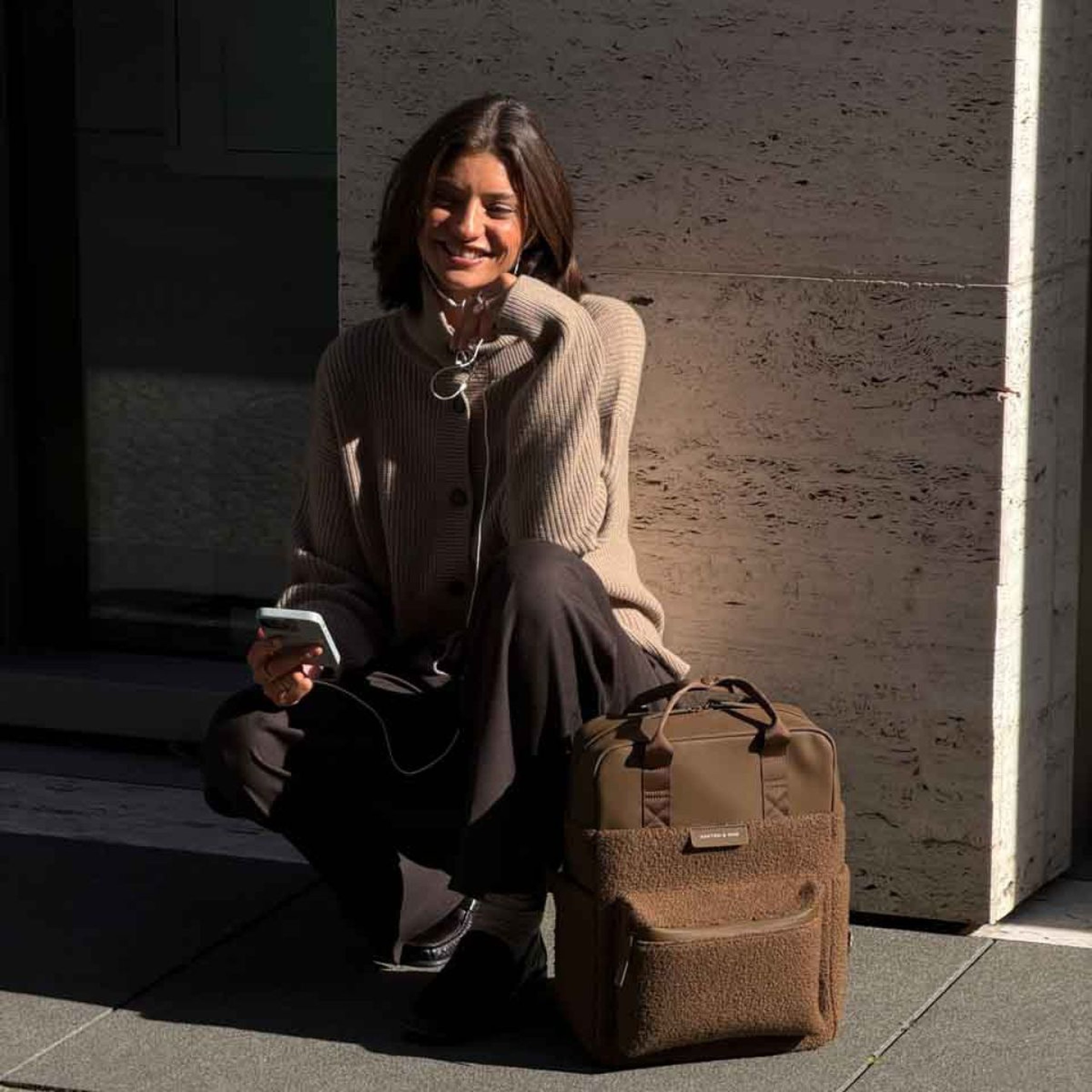 Lächelnde Frau in einer Strickjacke sitzt auf einer Stufe neben einem Kapten & Son Rucksack mit weichem Fleece und hält ein Smartphone.